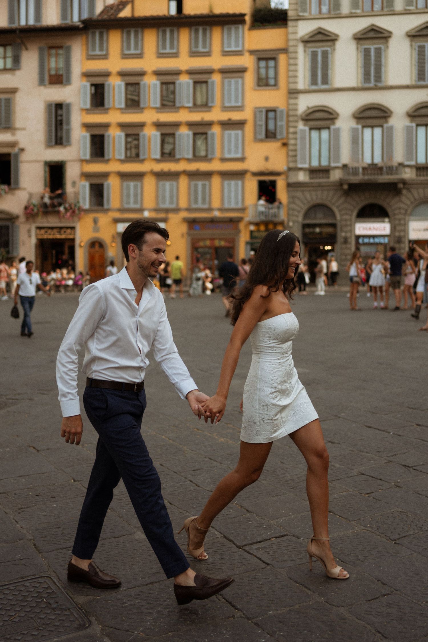 Couple in wedding attire holding hands walking through a sunlit Italian piazza in Florence.