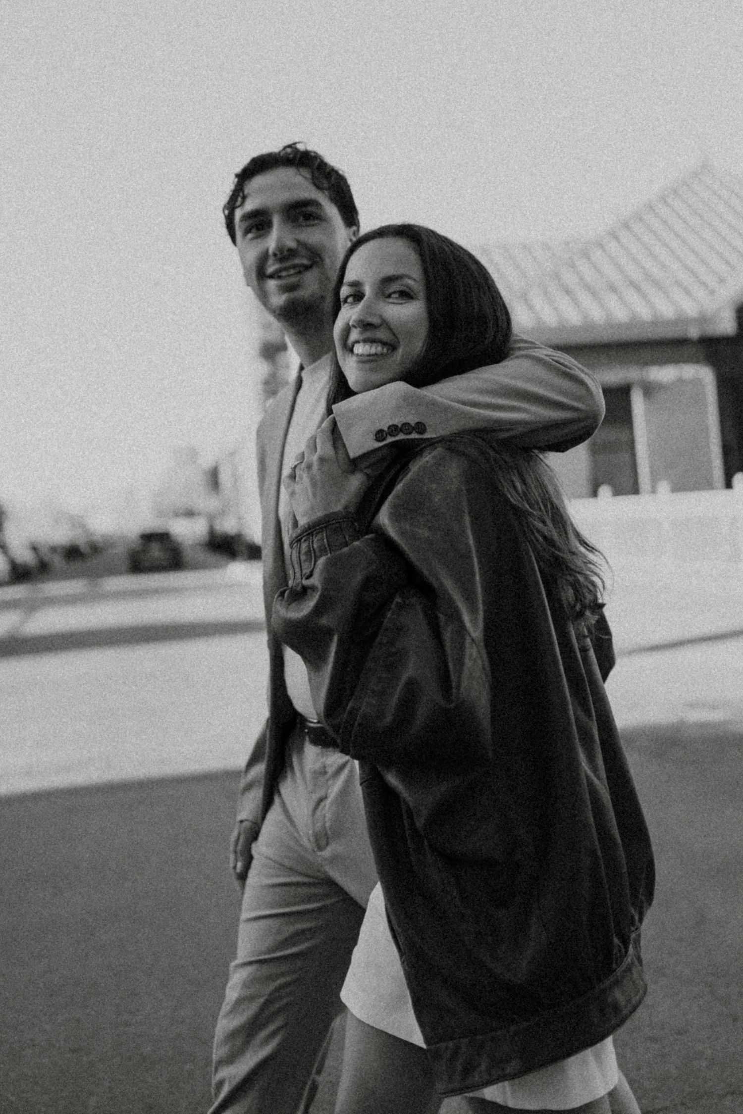 Black and white photo of a smiling couple walking together, woman hugging man from behind outdoors.