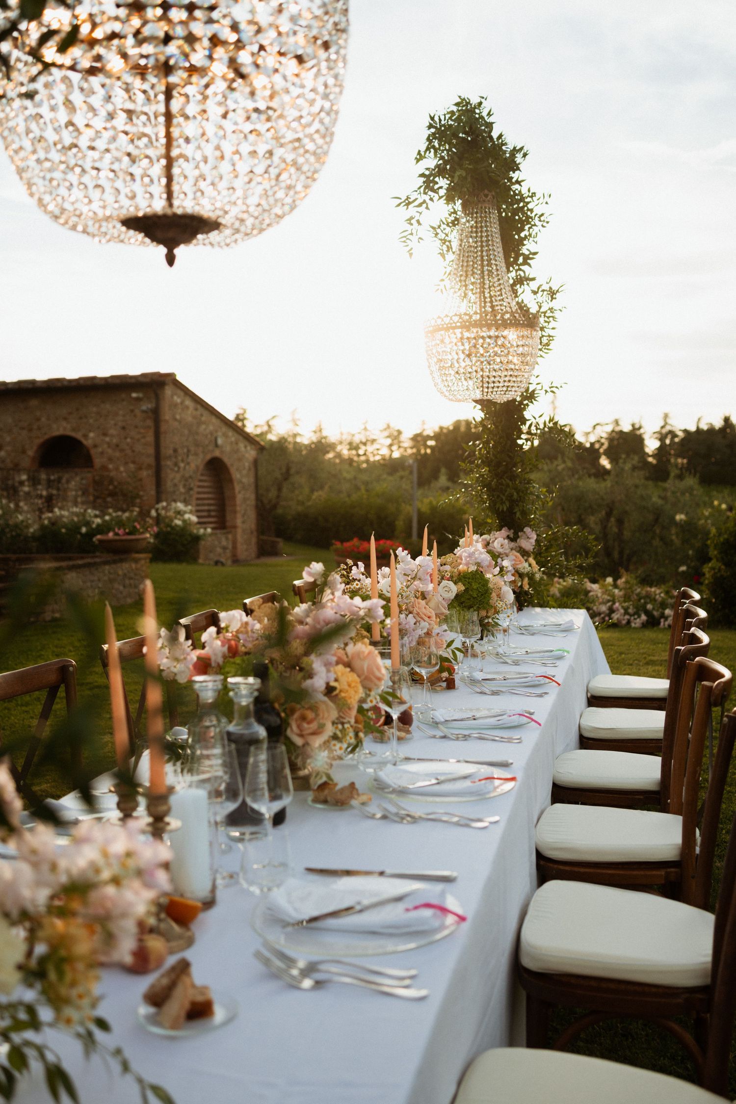 Elegant outdoor wedding reception table with crystal chandeliers, pink floral arrangements, and candlelight at golden hour.