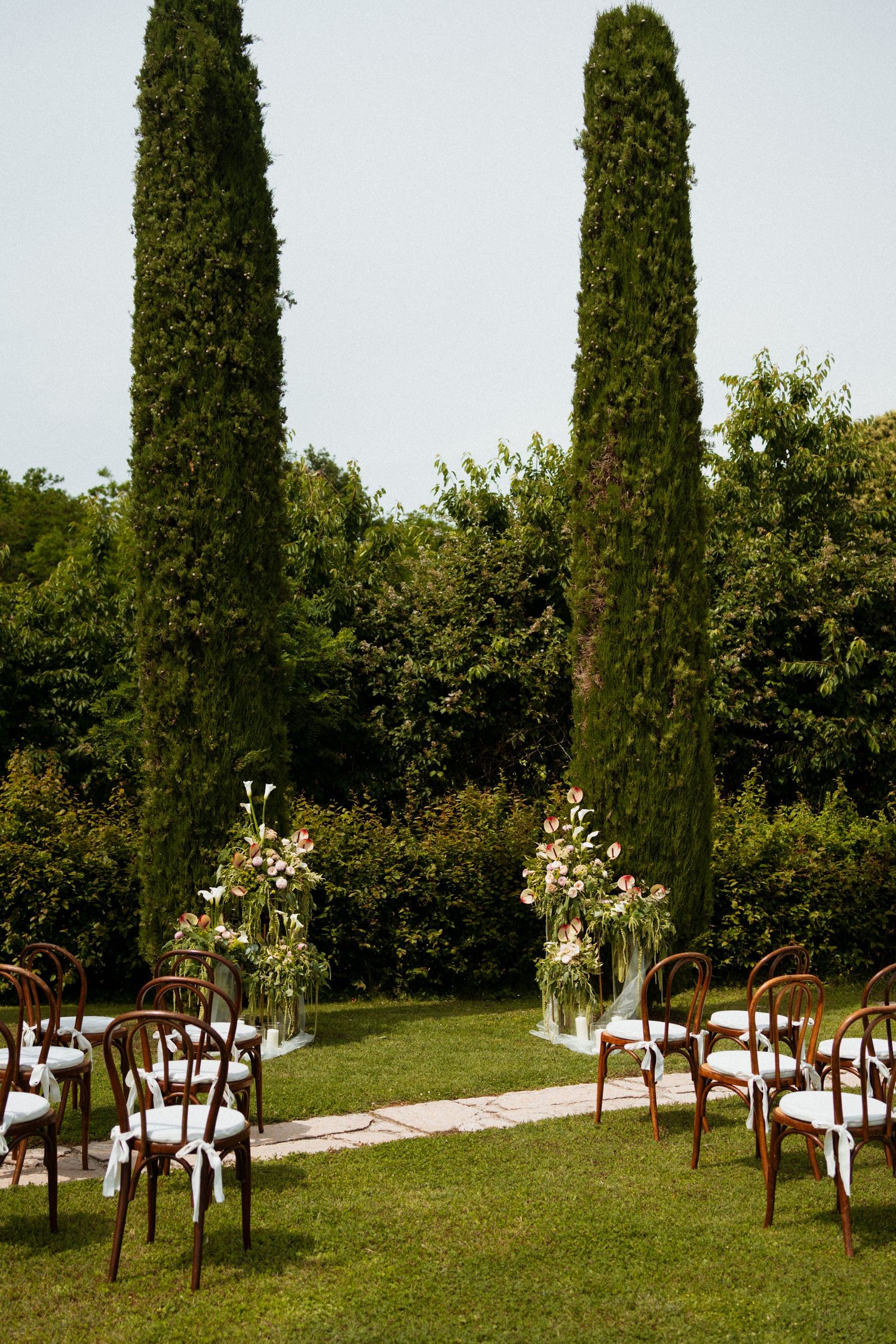Elegant outdoor wedding ceremony setup with wooden chairs, floral arrangements, and tall cypress trees in a lush garden.