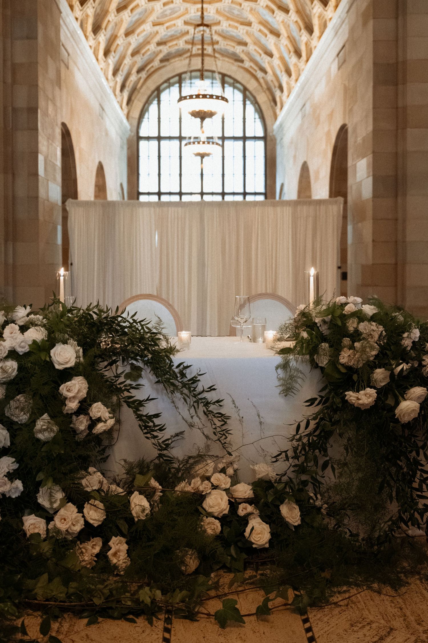 Elegant wedding ceremony setup with white floral wreaths, candles, and draped backdrop in a grand arched hall.