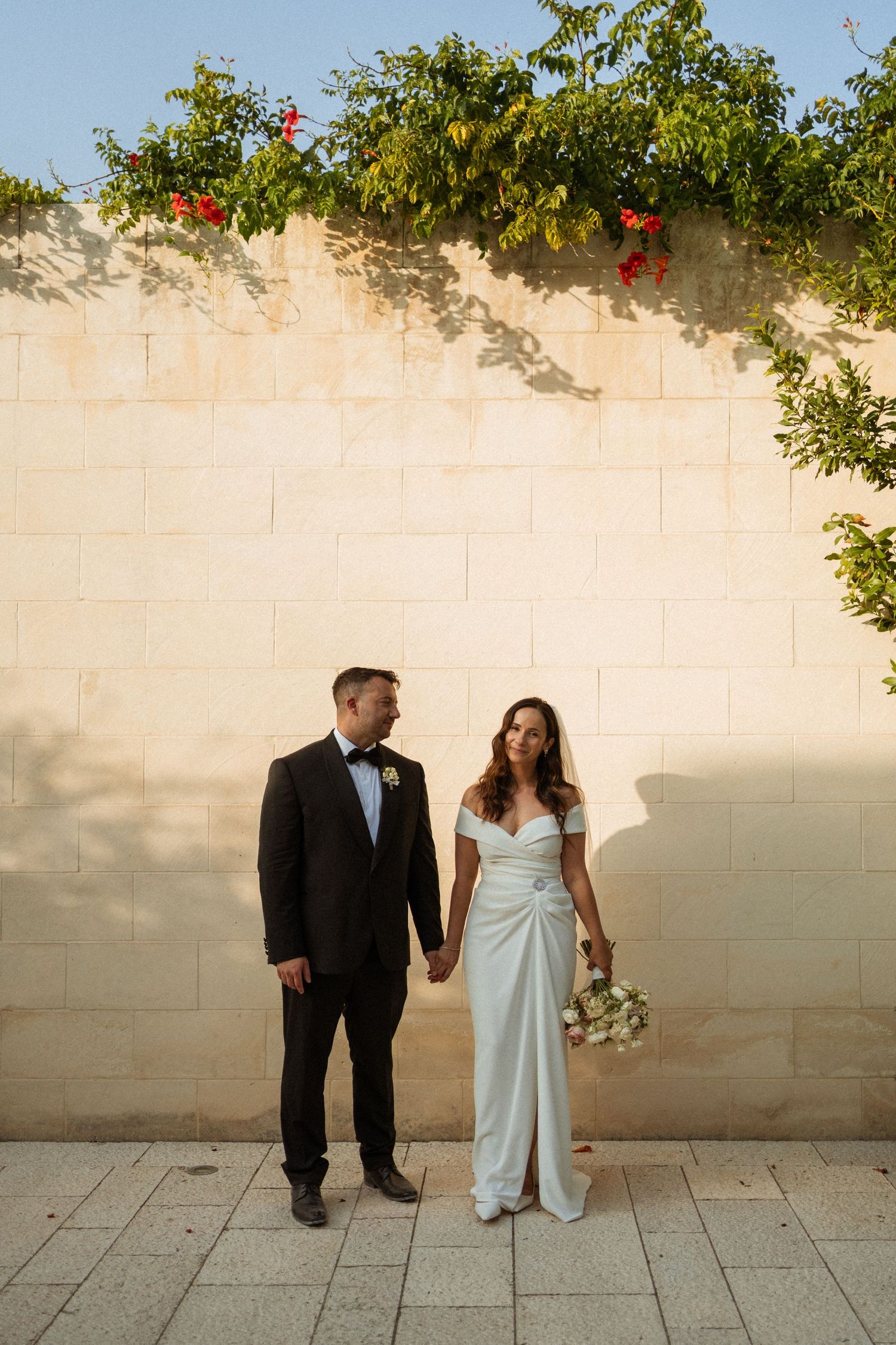 Couple in formal wedding attire pose against a light colored wall with green foliage and red flowers above.