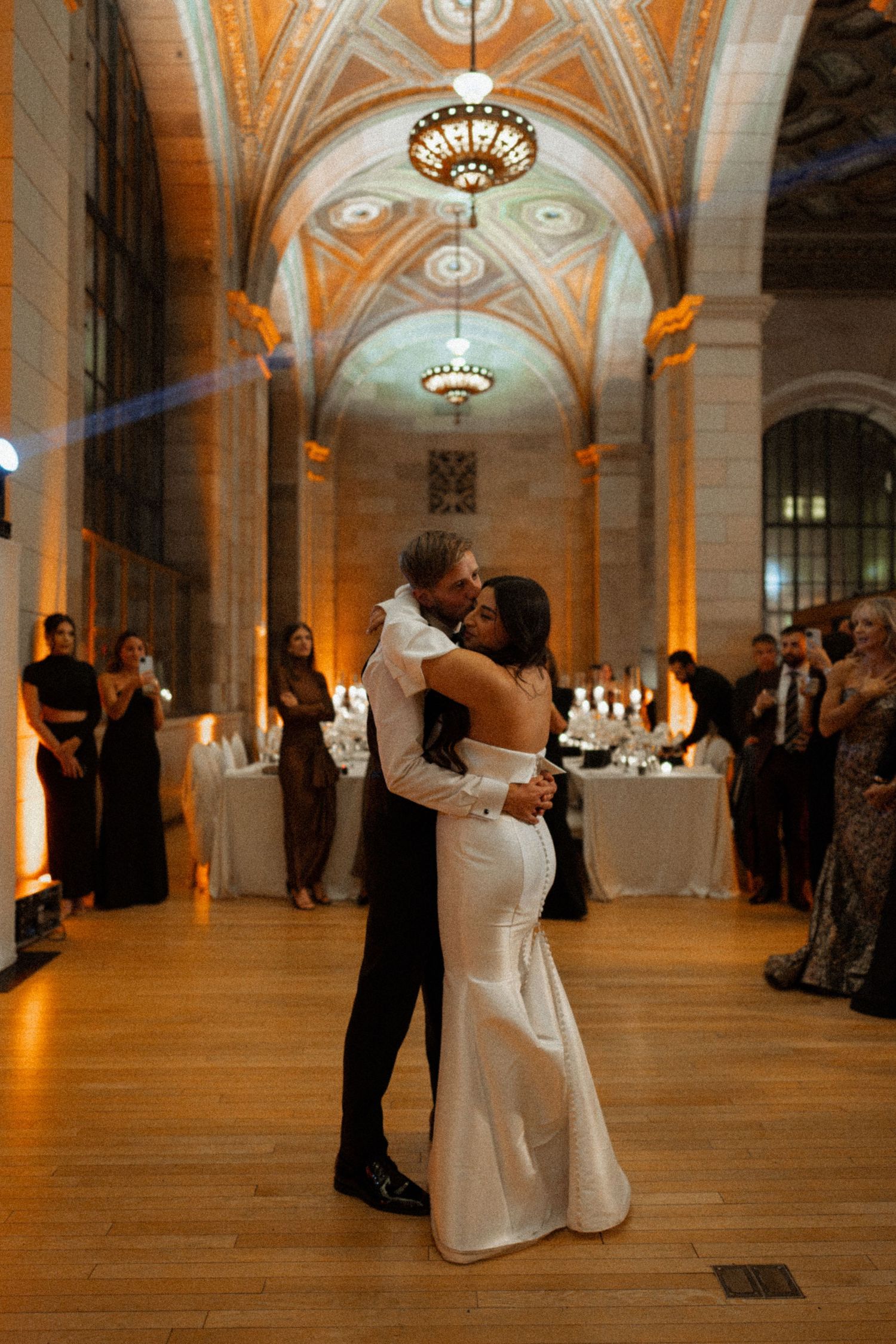A couple shares their first dance in an elegant ballroom with vaulted ceilings and ornate chandeliers.