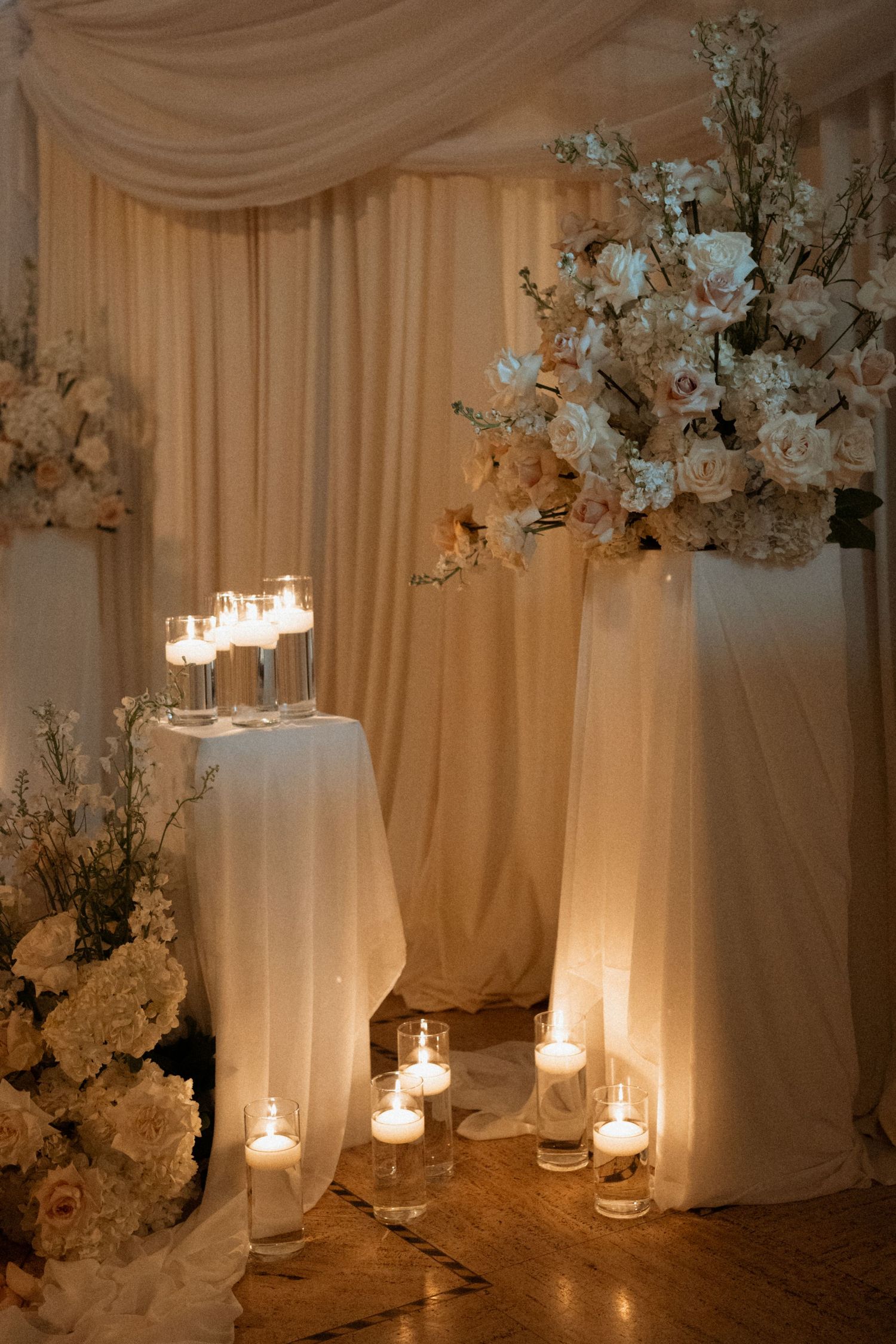 Elegant wedding reception decor features white floral arrangements and glowing candles against draped fabric backdrops.