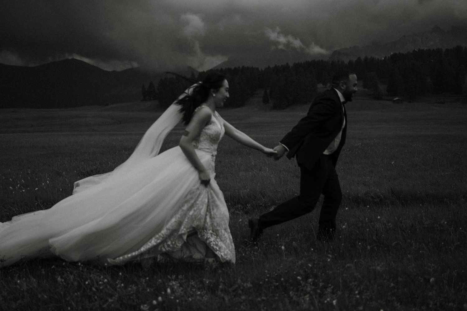 Black and white photo of a wedding couple running playfully through a meadow.
