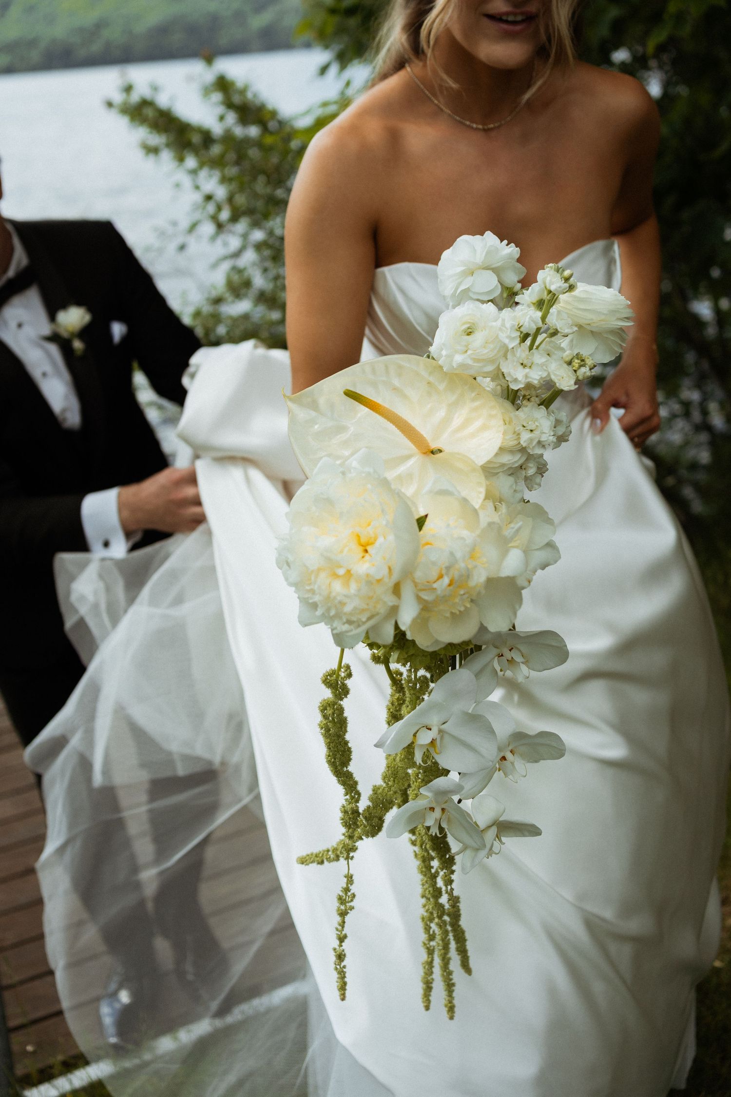 Bride in strapless white gown holds cascading bouquet of white anthuriums, peonies, and orchids near a lakeside.
