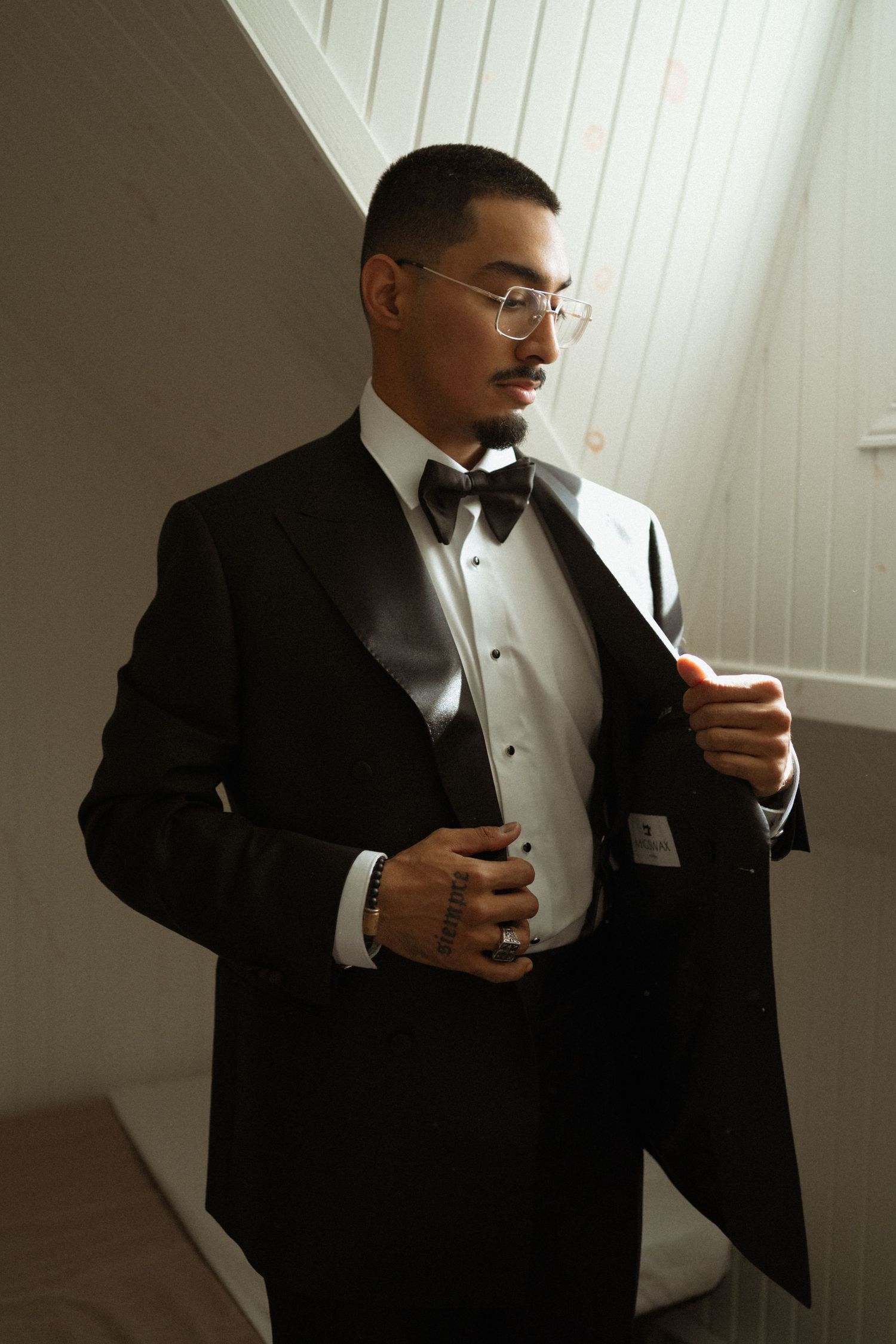 A person in a black suit takes a sip from a coffee cup while standing against a white wall.