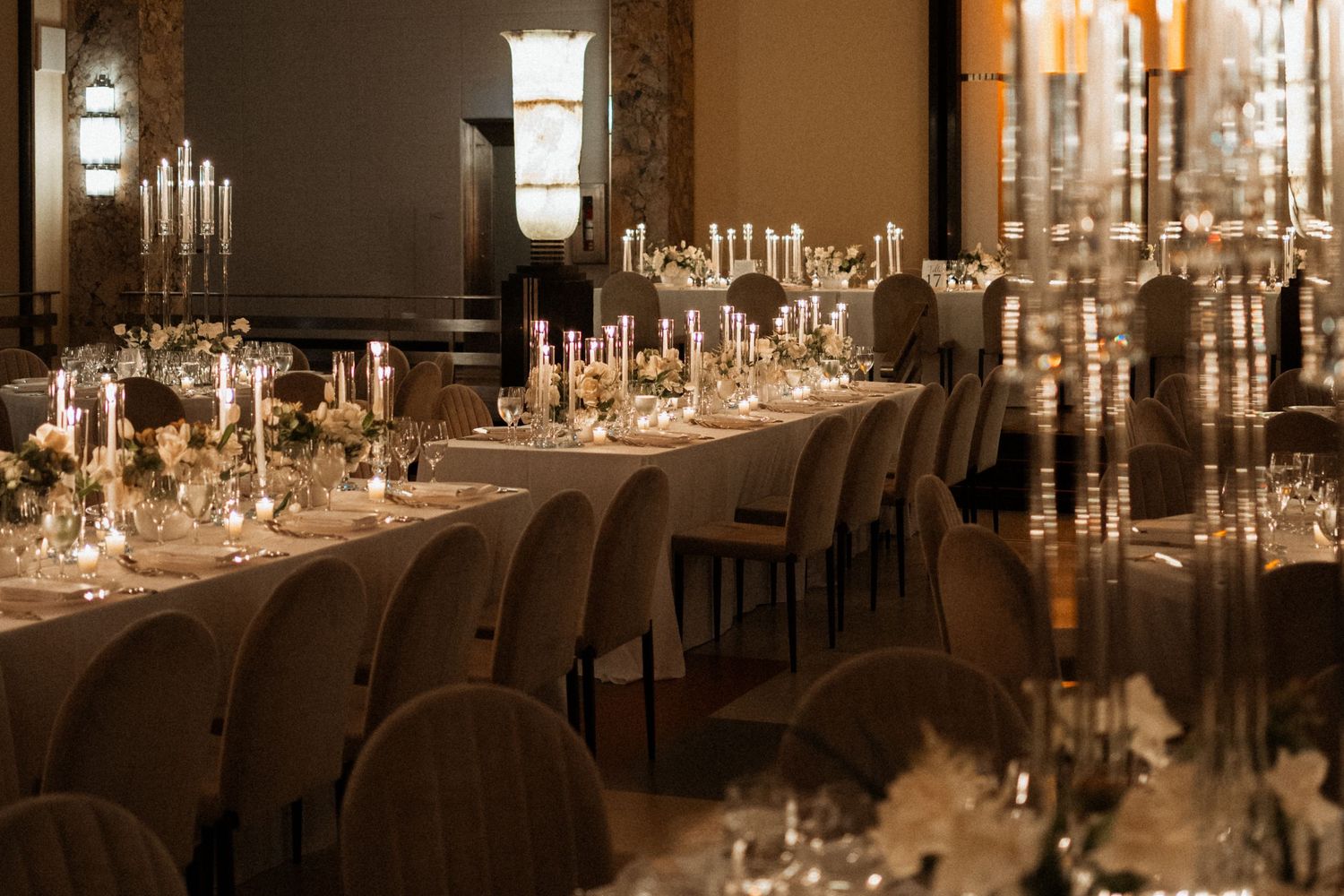 Elegant wedding reception venue with crystal chandeliers and long tables set with glowing candles and white floral centerpieces.