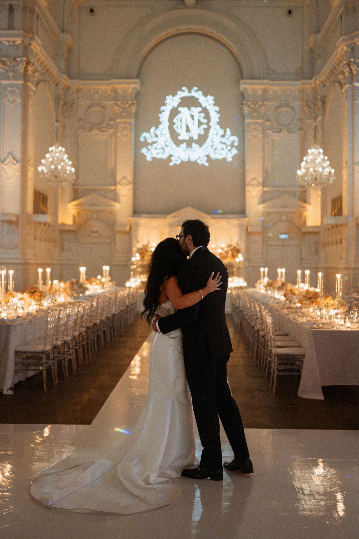 Elegant wedding couple dancing in a grand ballroom with candlelit tables and ornate decor creating a romantic atmosphere.