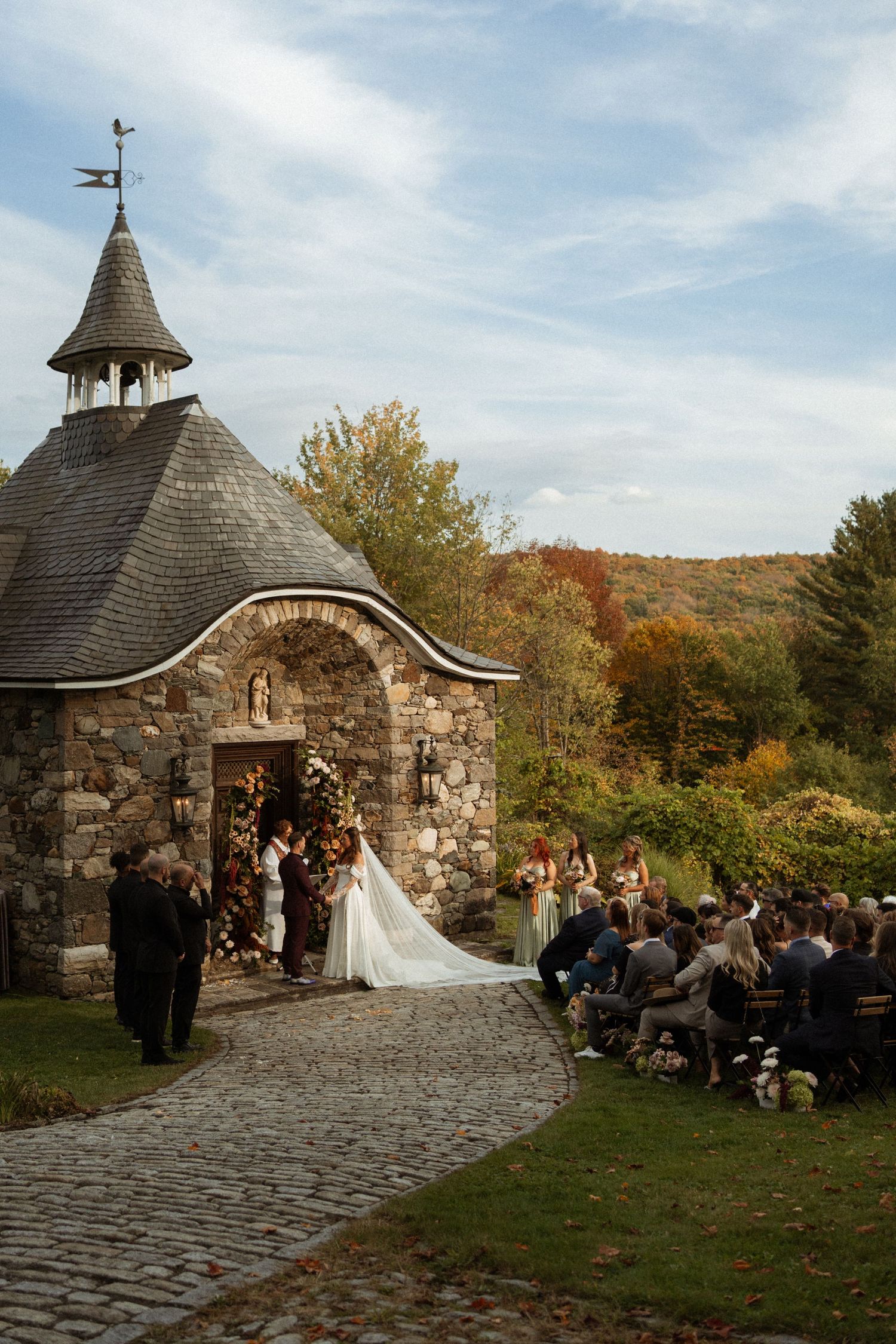 Stone chapel wedding ceremony venue surrounded by autumn trees and gathered guests.