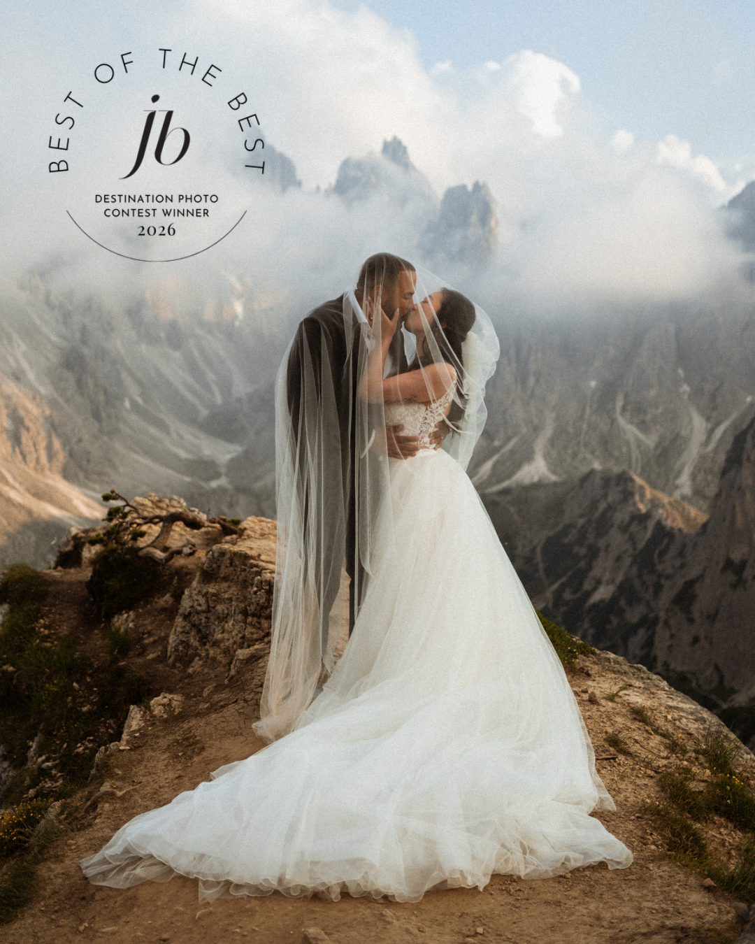 Bride and groom kissing on a rocky mountain peak in the Dolomites, with dramatic clouds and rugged peaks behind them.