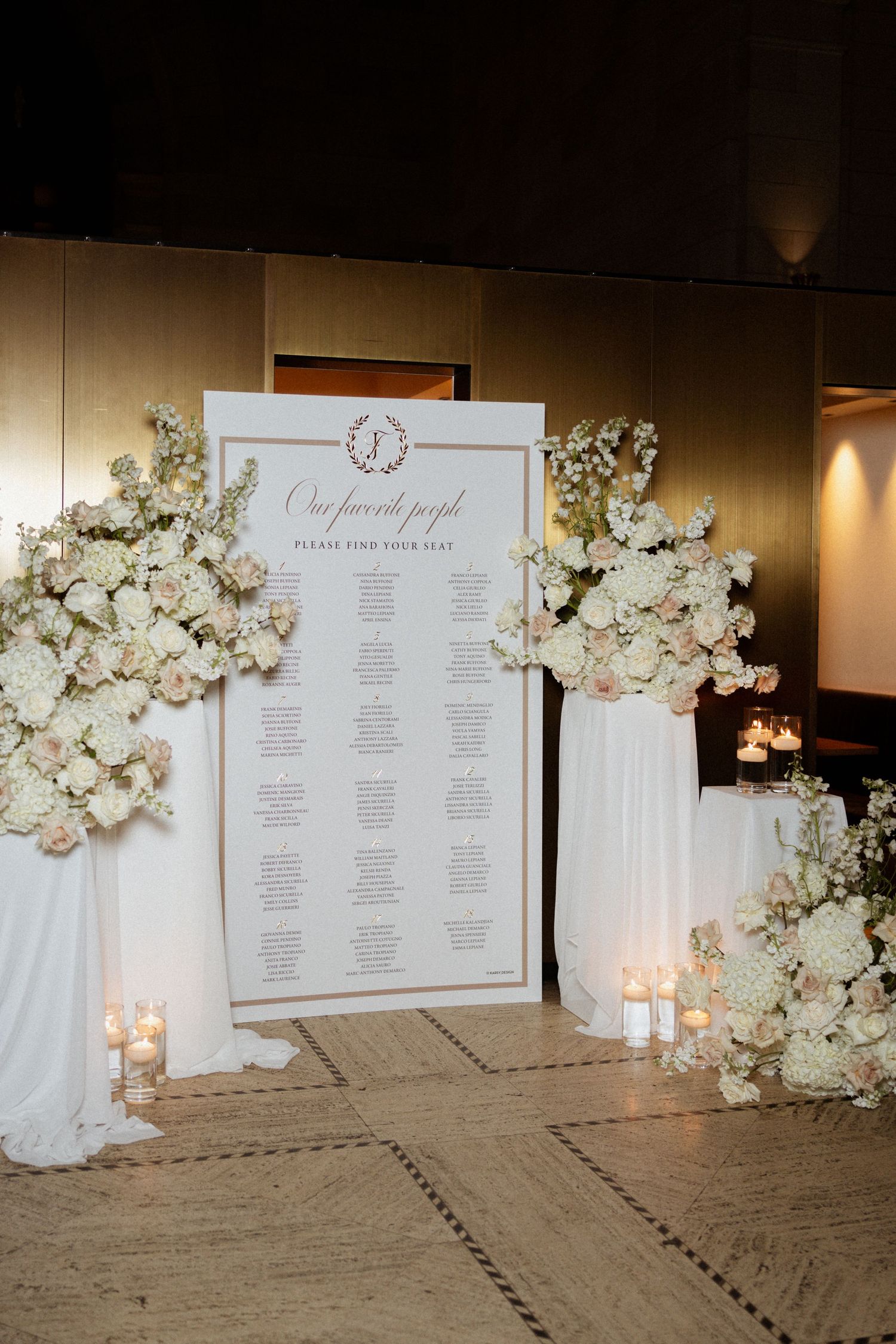 Elegant wedding seating chart display with white floral arrangements, candles, and draped fabric at a formal reception venue.
