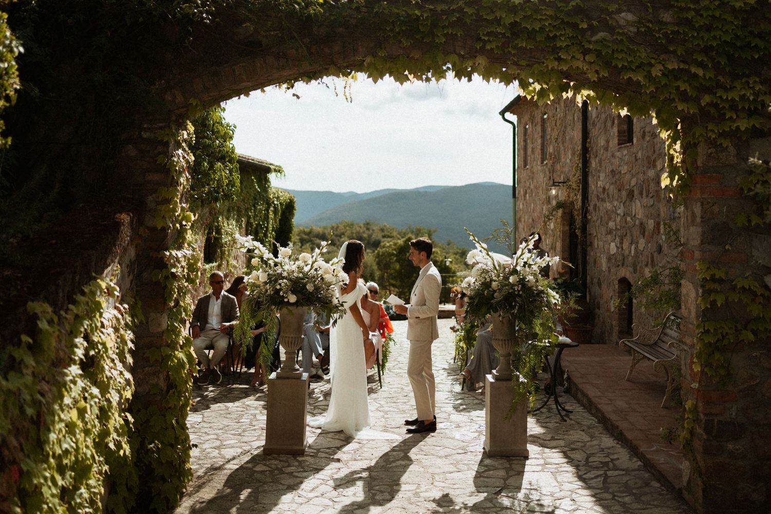 A wedding couple shares a romantic moment in a sun-drenched stone archway surrounded by lush greenery.
