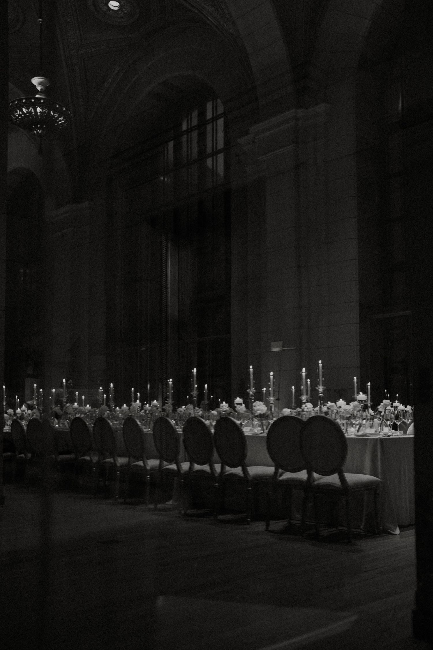 Black and white photo of an elegant candlelit banquet hall with long dining tables and ornate chairs in a grand venue.