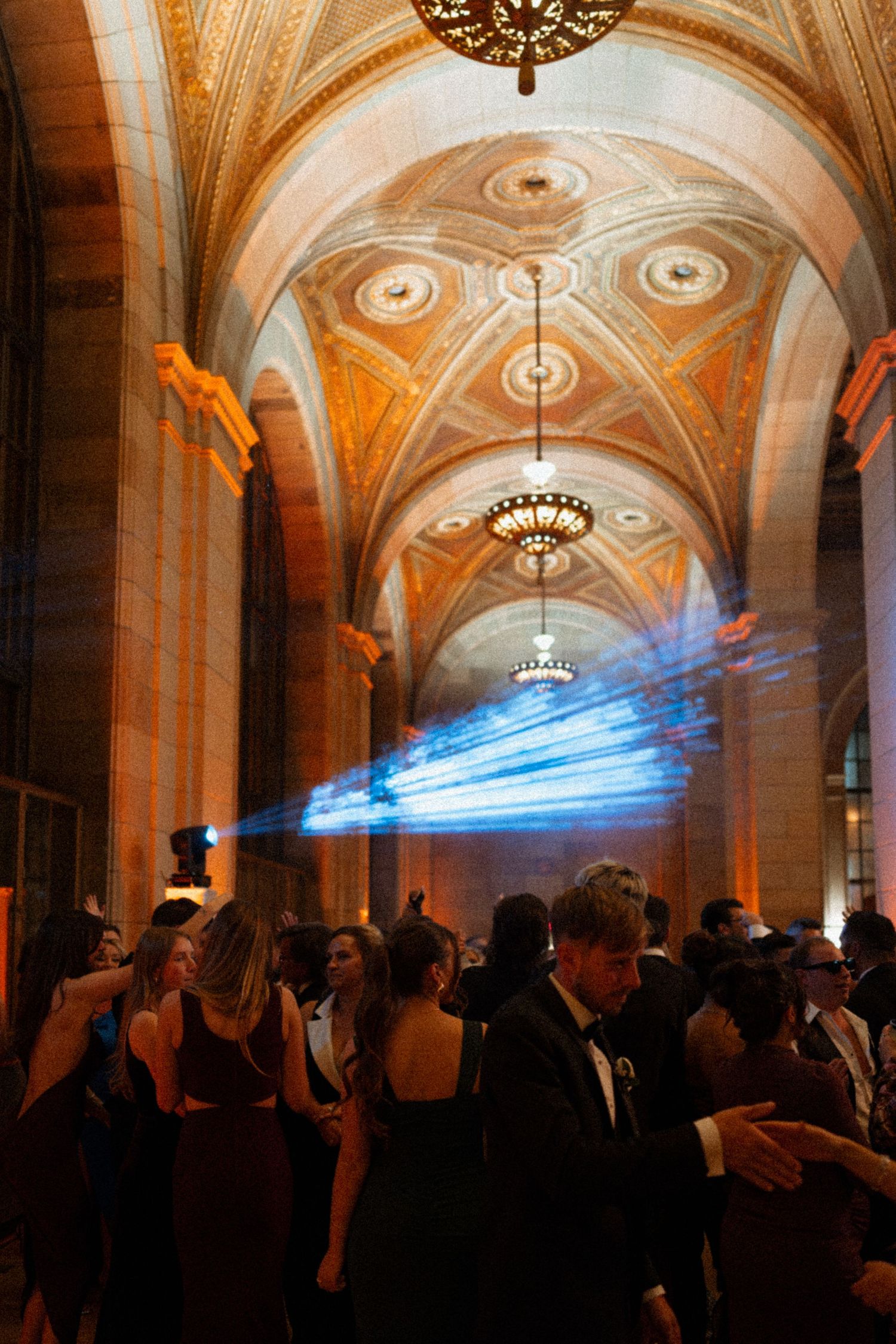 Elegant wedding reception in ornate vaulted hall with blue laser lights, guests dancing in formal attire under golden arched ceilings.