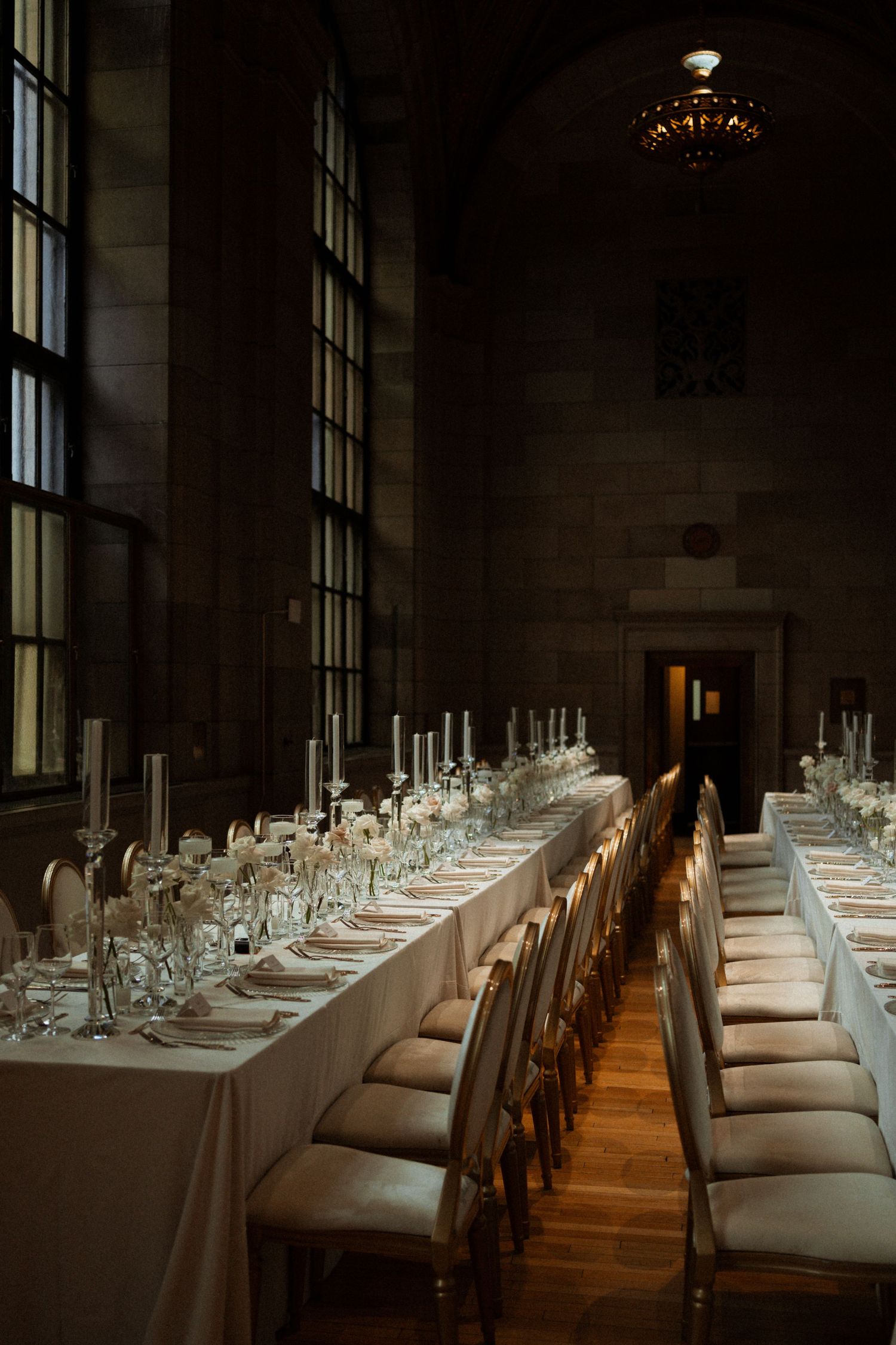 Long banquet table set with elegant white linens and place settings in a dimly lit room with dramatic window lighting.