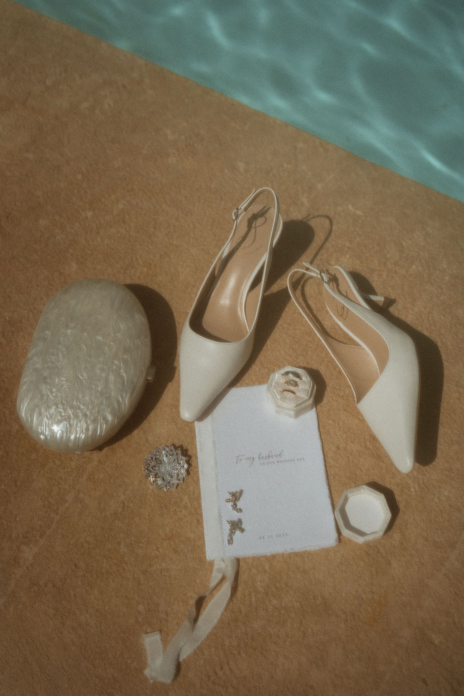 Wedding accessories including white shoes, jewelry, and a small purse arranged by a poolside.