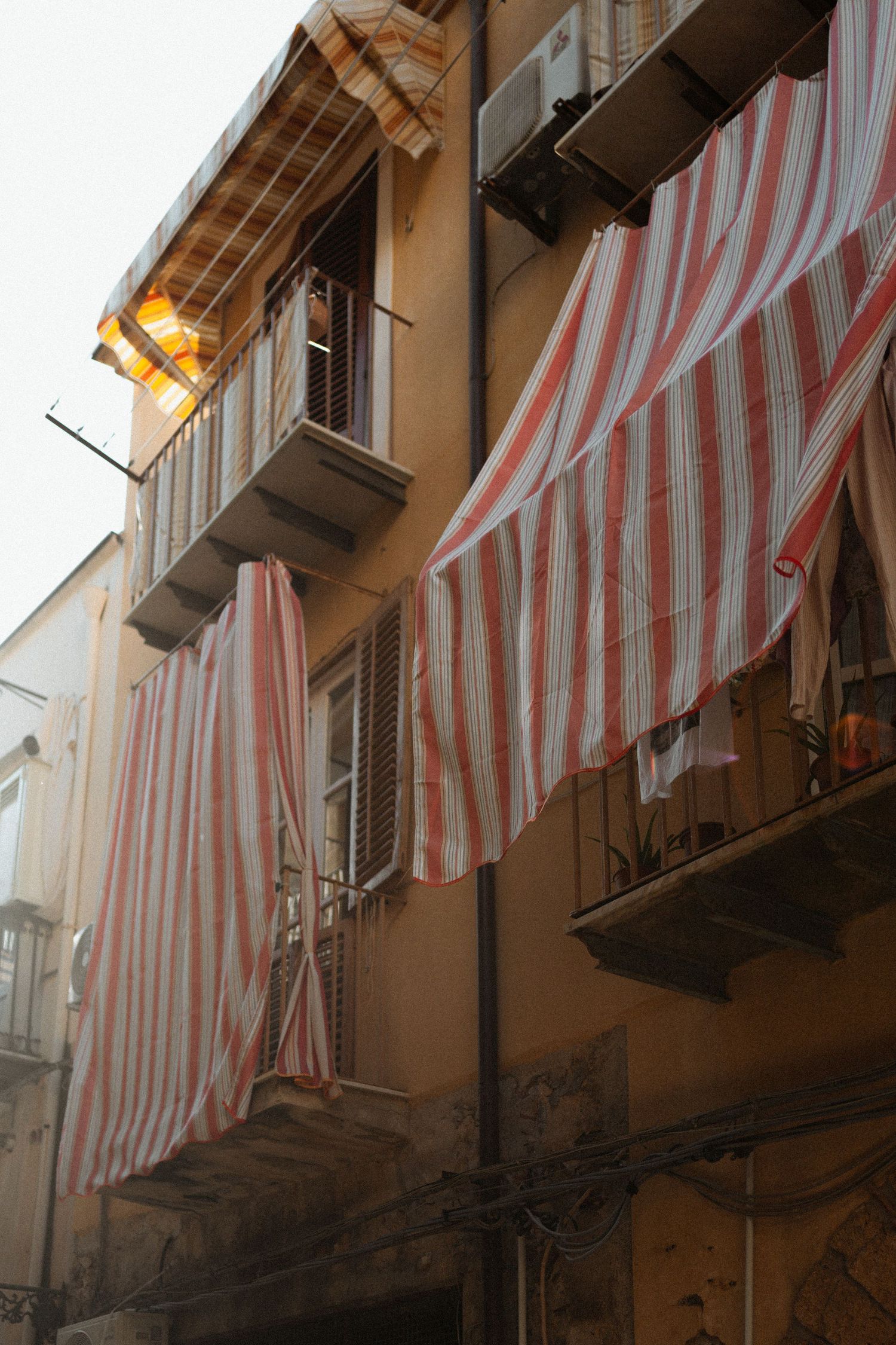 Red and white striped awnings hang from balconies on an old Italian apartment building facade in warm sunlight.