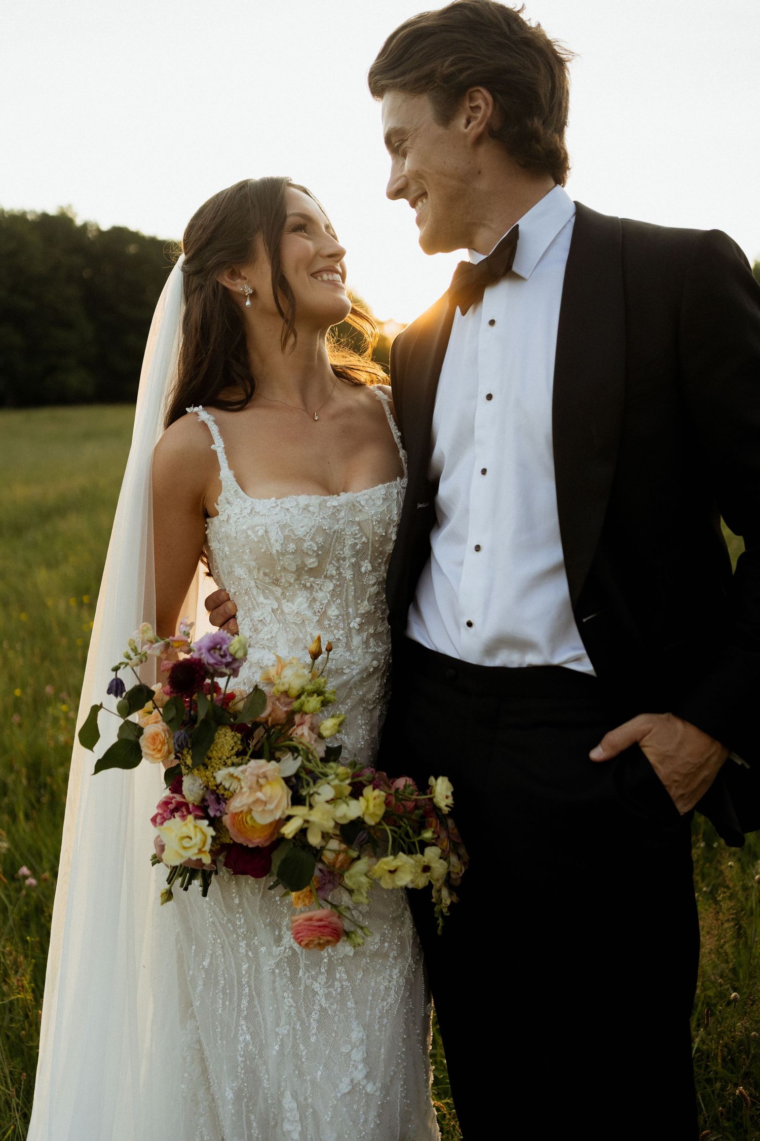 A bride and groom smile at each other in a sunlit field, she holds a colorful bouquet wearing a lace gown with veil.