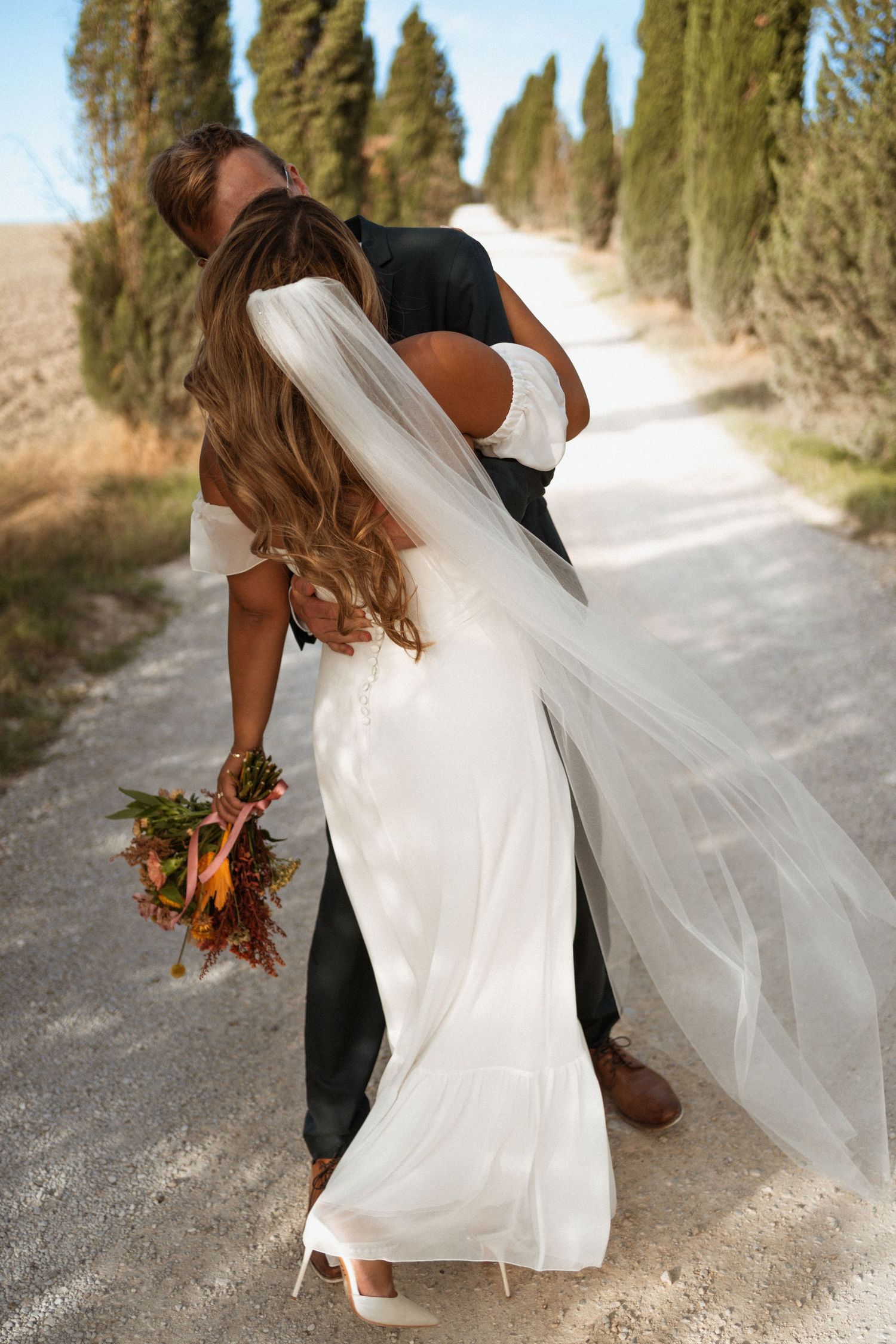 A romantic wedding couple walking along a tree-lined path as the bride's white veil flows elegantly in the breeze.