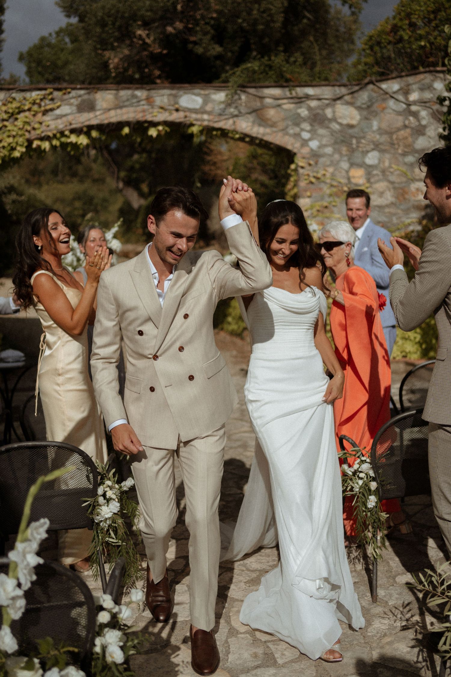 Joyful newlyweds walk hand in hand at outdoor wedding ceremony, groom in beige suit, bride in white gown, guests cheering.