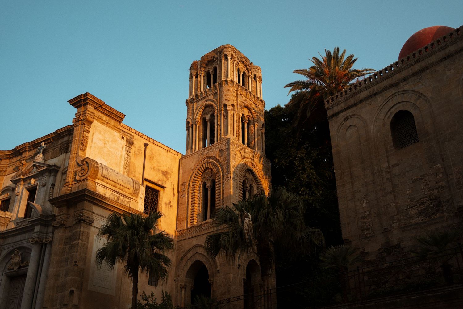 Historic Mediterranean church with palm trees and stone architecture illuminated by warm sunset light.