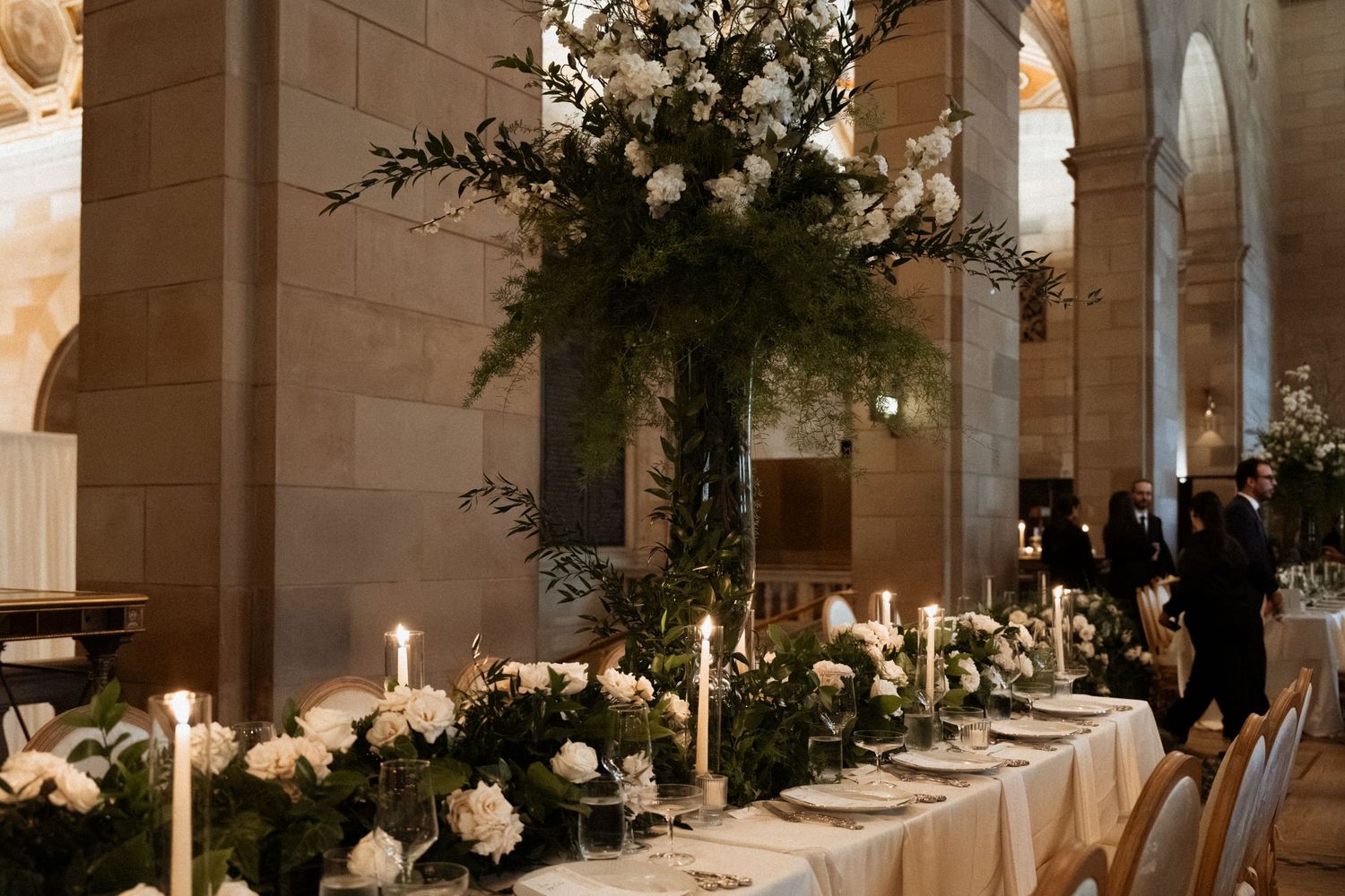 Elegant wedding reception table with tall floral arrangements and glowing candles in a grand stone venue.
