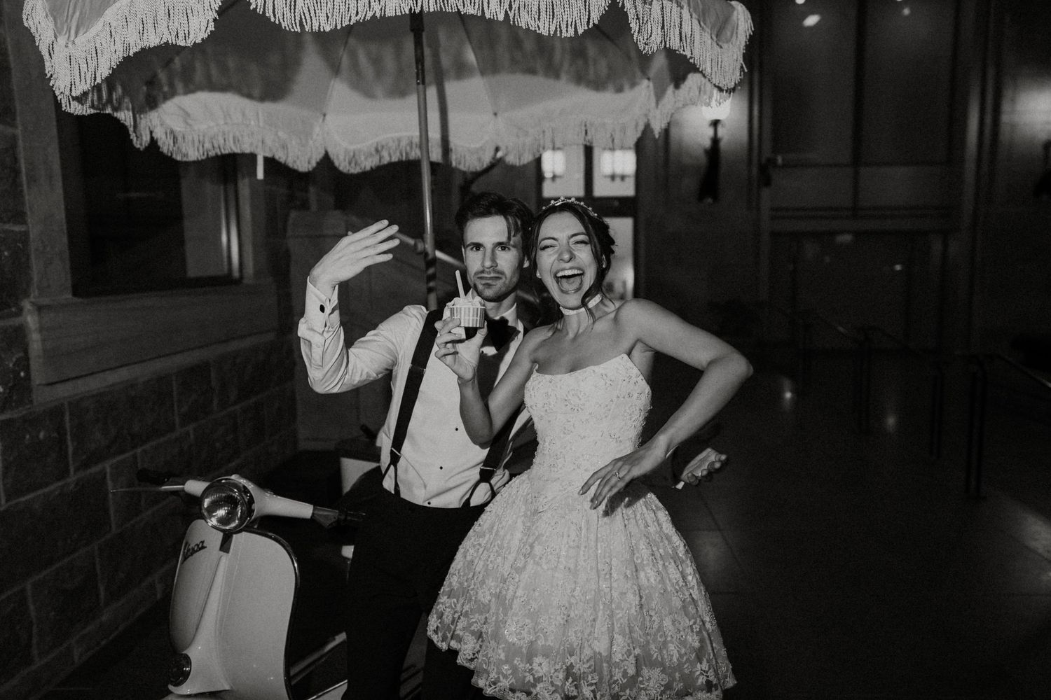 Black and white photo of a couple at a vintage-style wedding celebration posing with parasols under decorative lighting.