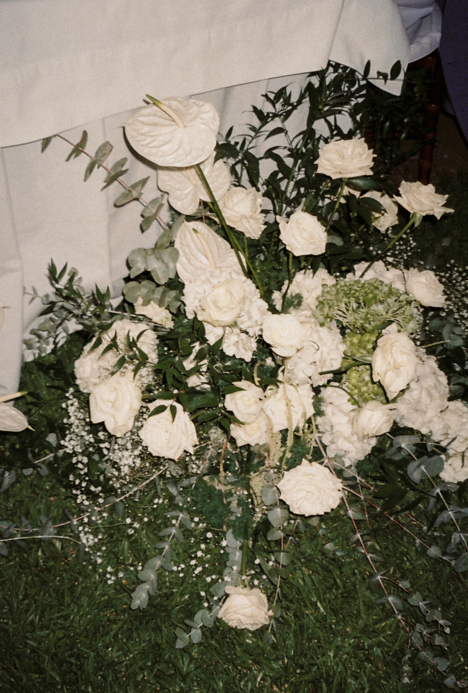 A sequence of white roses and greenery arranged against a wall in dim lighting creates a romantic garden atmosphere.