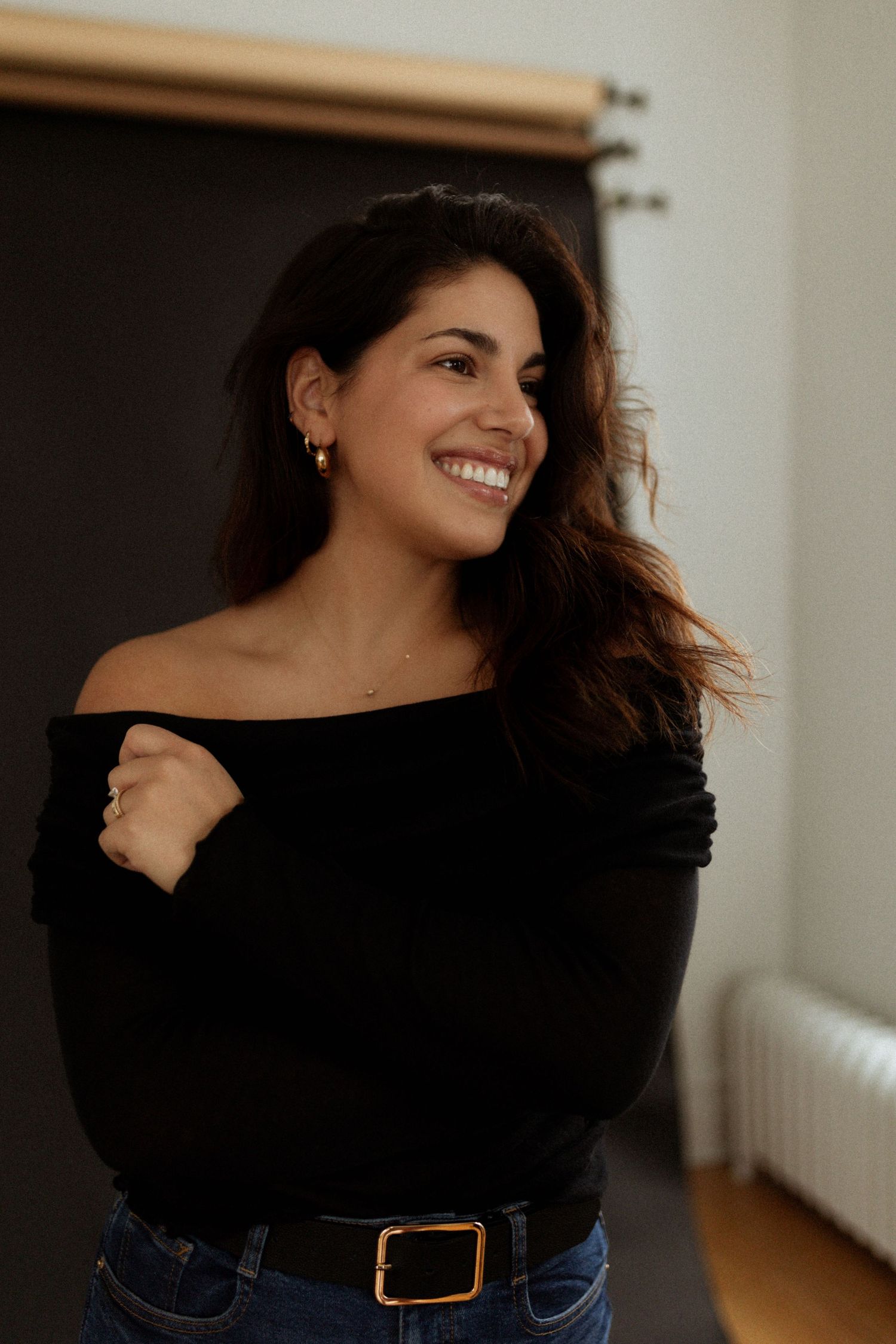 Woman in black off-shoulder top smiling and posing against light interior background.