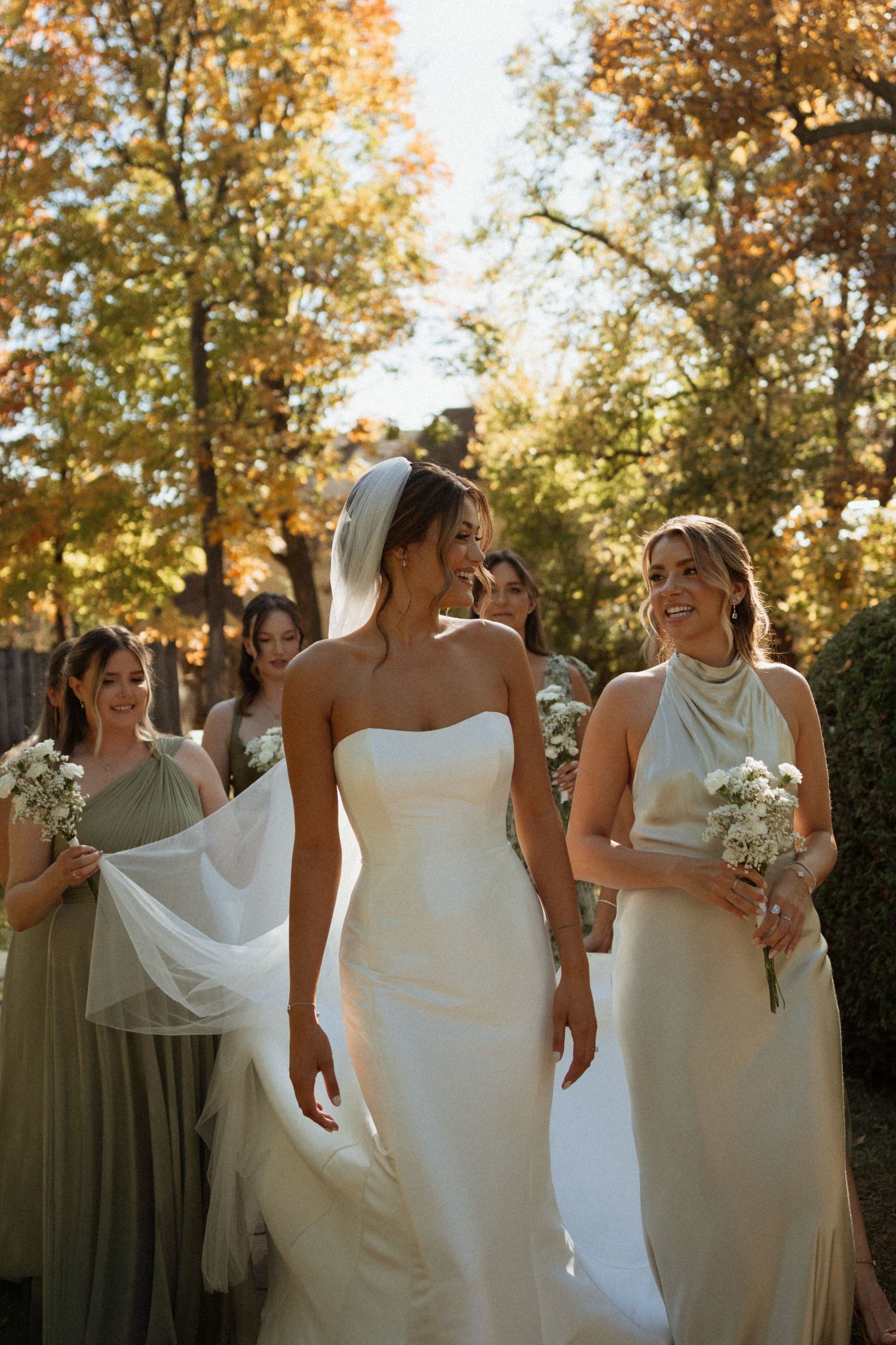 Wedding party laughs together outdoors in autumn with bridesmaids wearing white dresses and olive green gowns.