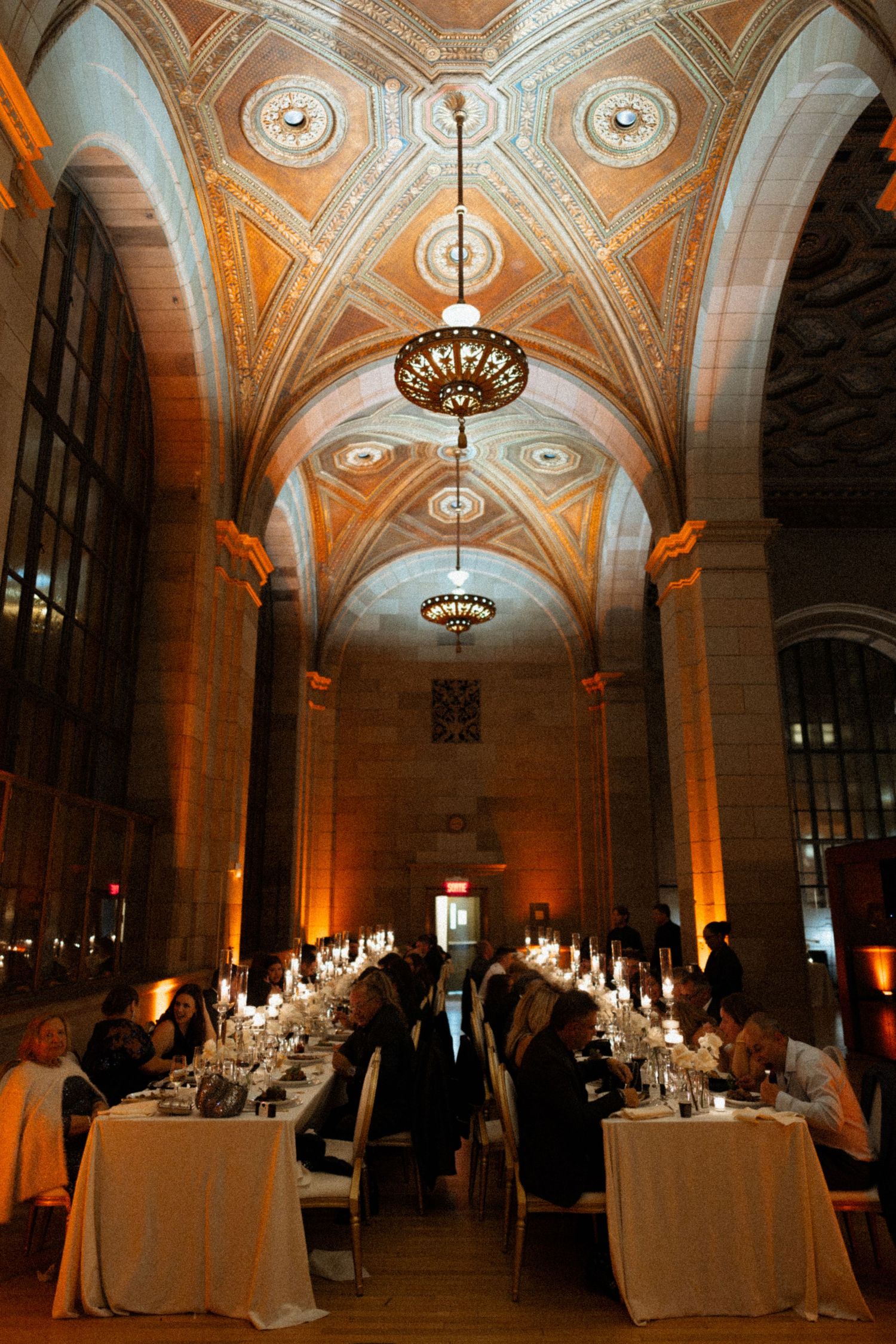 Elegant candlelit dinner in ornate vaulted hall with gilded ceilings, long banquet tables, and warm amber lighting.