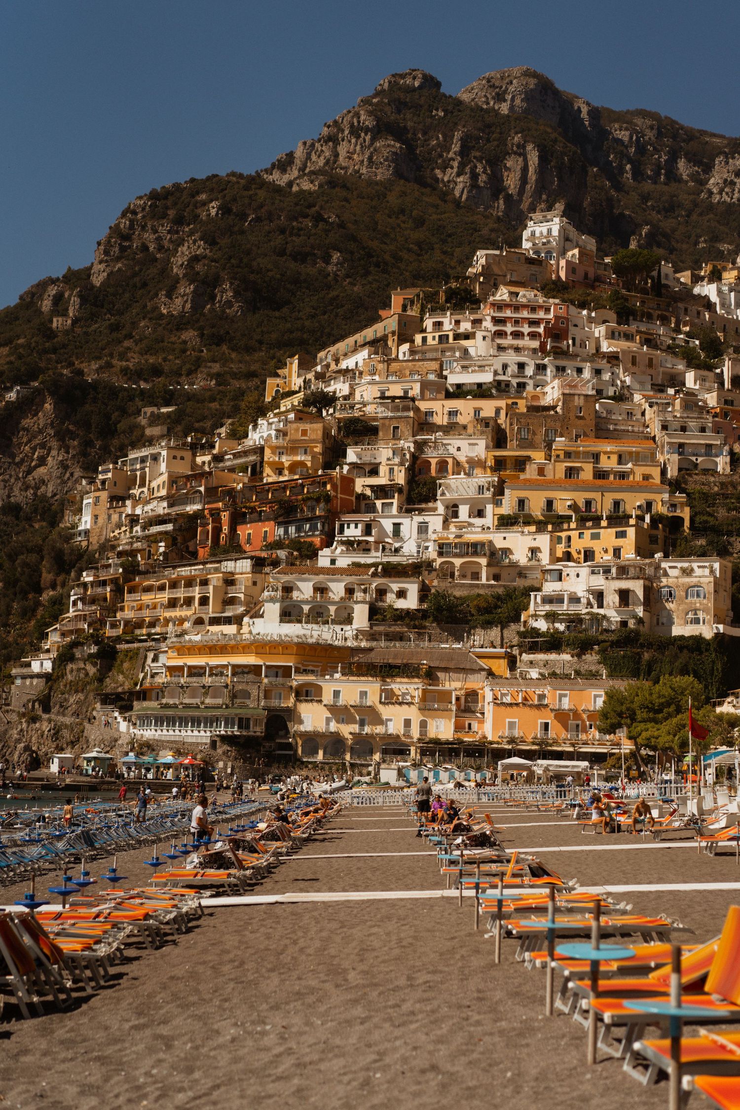 Orange beach chairs line the sandy shore of Positano, Italy with its iconic cliffside buildings cascading down the mountainside.