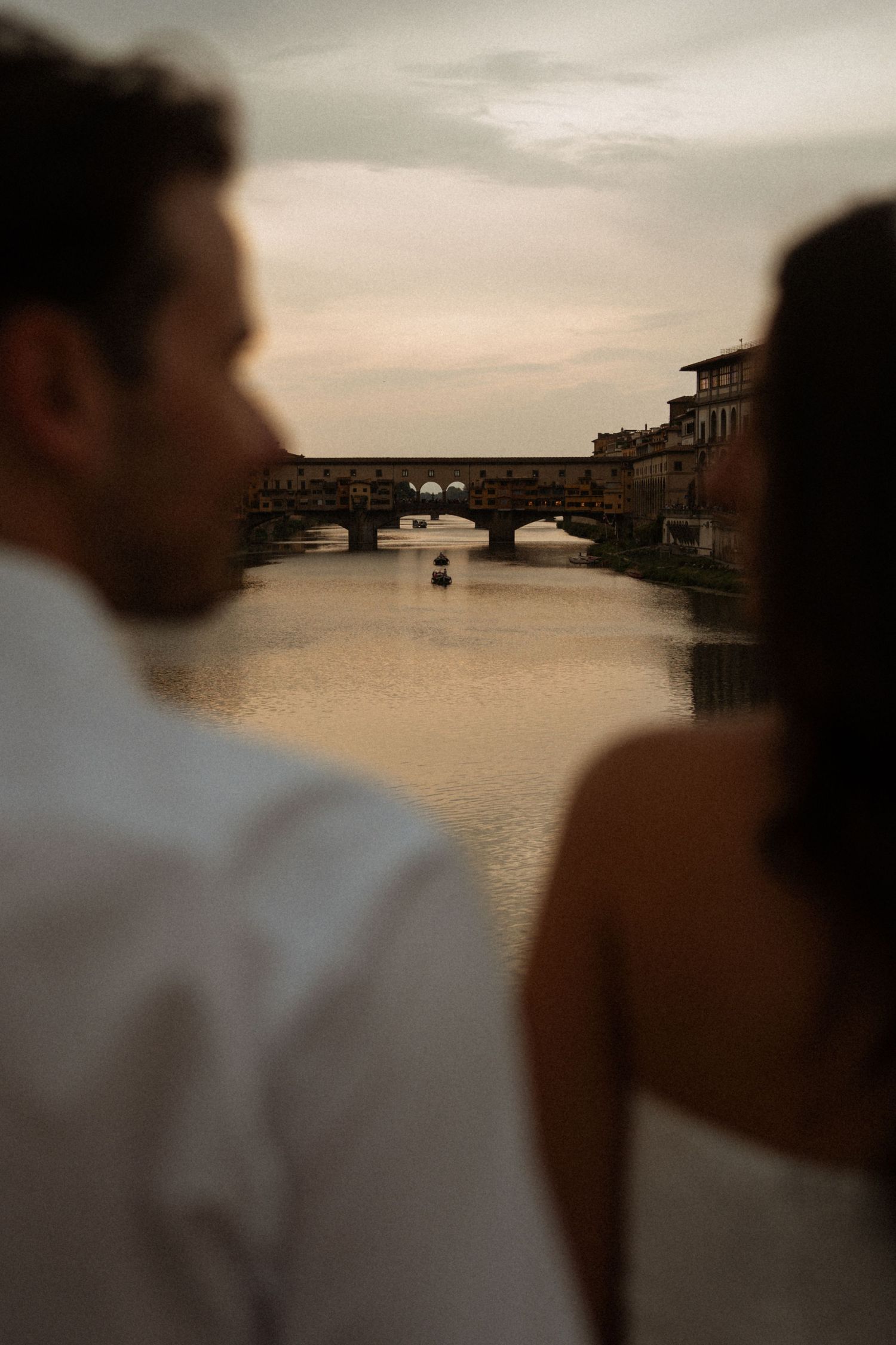 Couple in wedding attire gazing at Ponte Vecchio bridge over the Arno River in Florence, Italy at sunset.