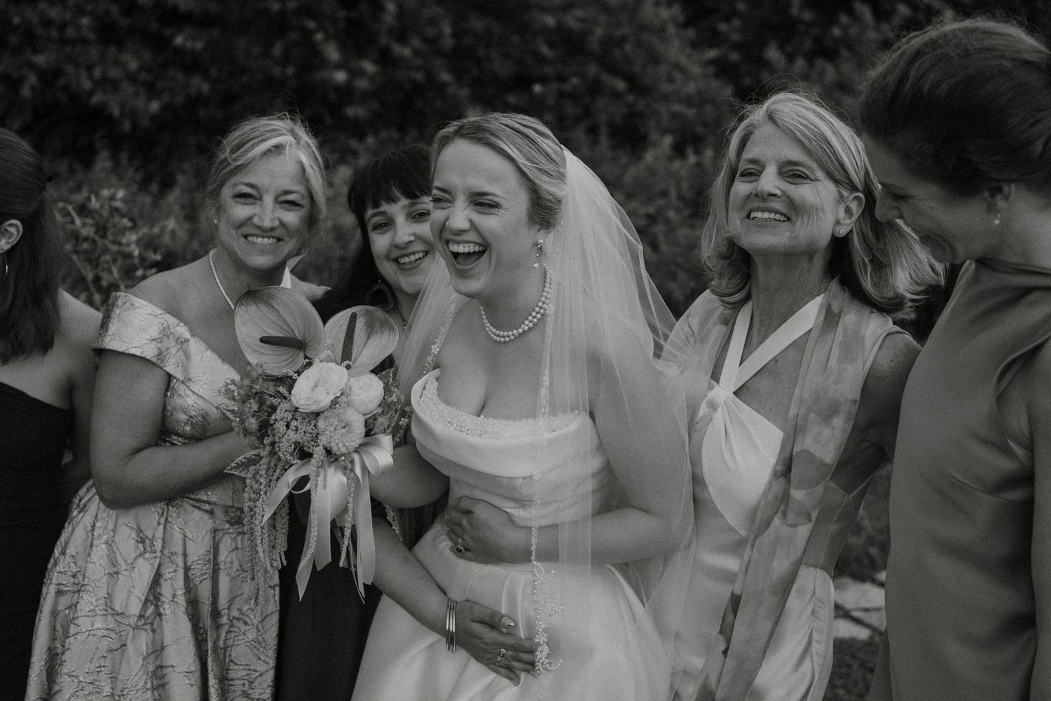 Black and white photo of a laughing bride in veil and gown surrounded by smiling women at an outdoor wedding celebration.