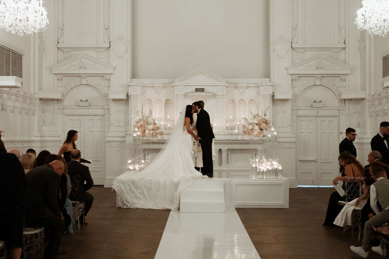 A couple shares their first kiss during an elegant wedding ceremony in a grand white room with crystal chandeliers.
