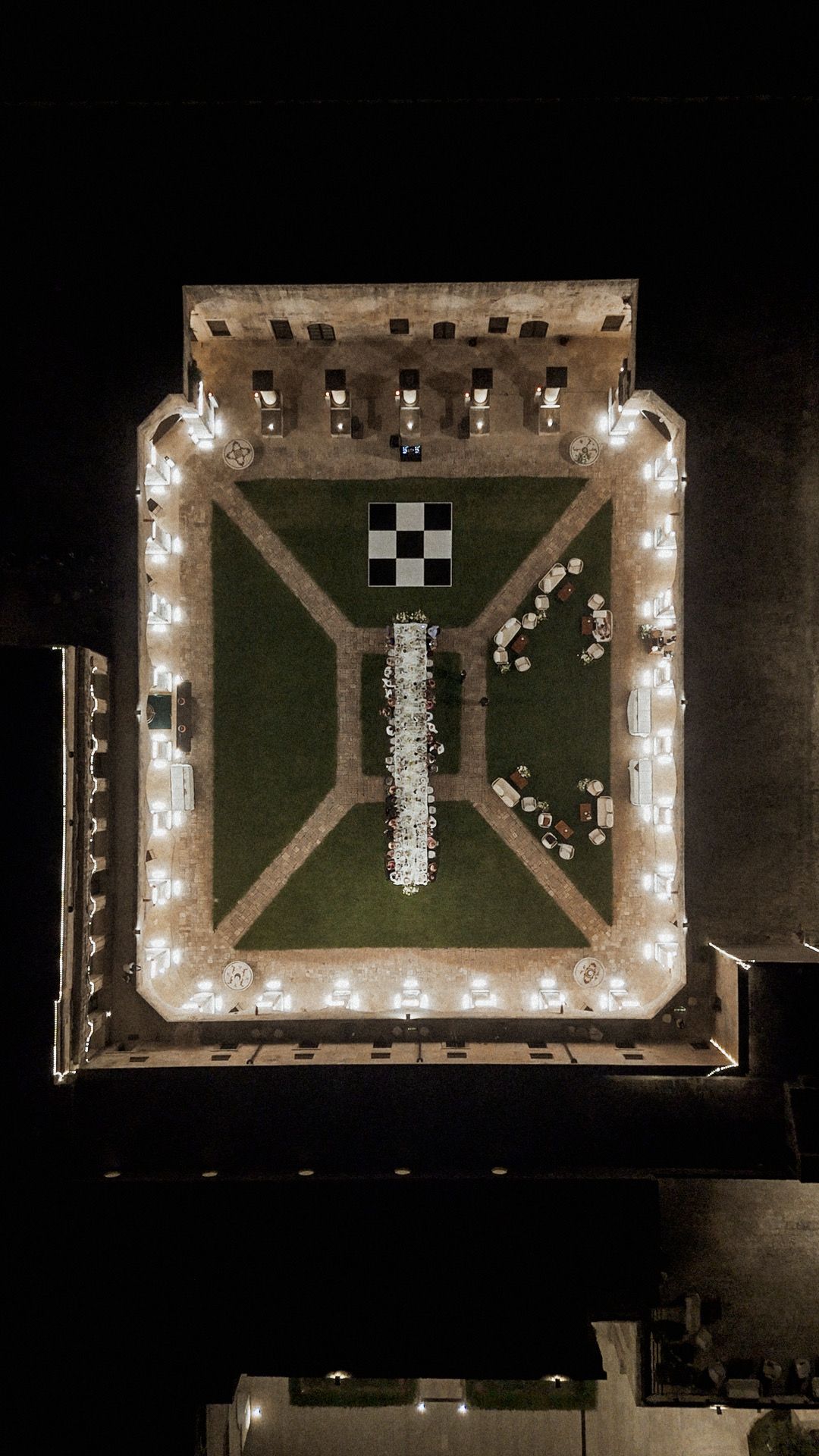 Aerial night view of an illuminated outdoor venue with a checkerboard pattern and seating arrangement on green lawn.