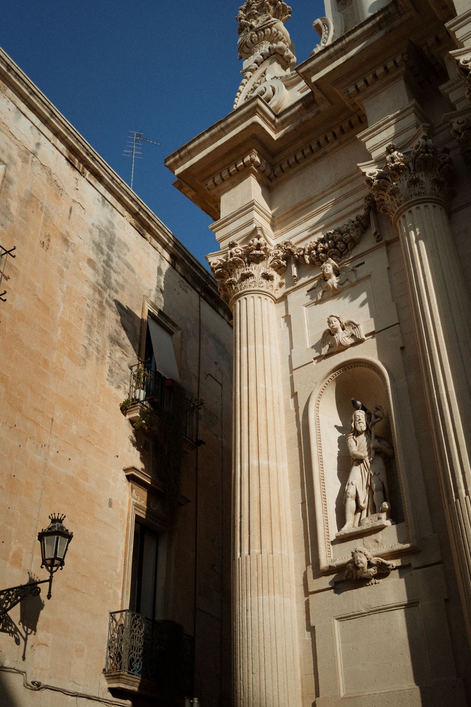 Ancient stone architecture with ornate Corinthian columns and religious statue in alcove against bright blue sky in Italy.