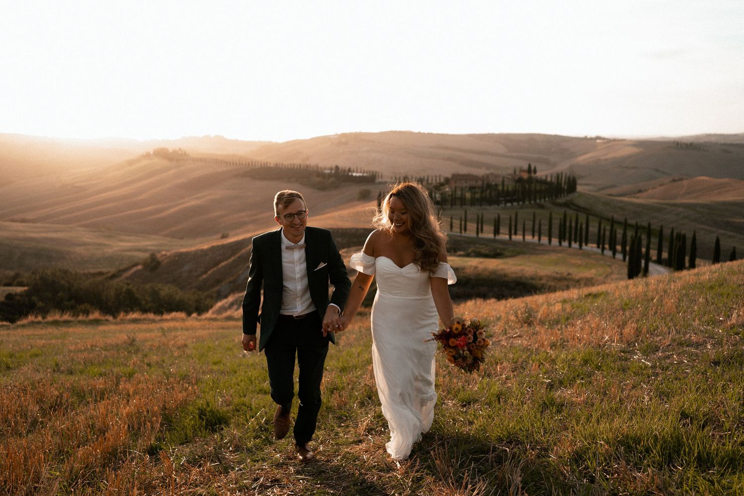 A couple walks hand in hand through the rolling hills of Tuscany with cypress trees lining the horizon at sunset.