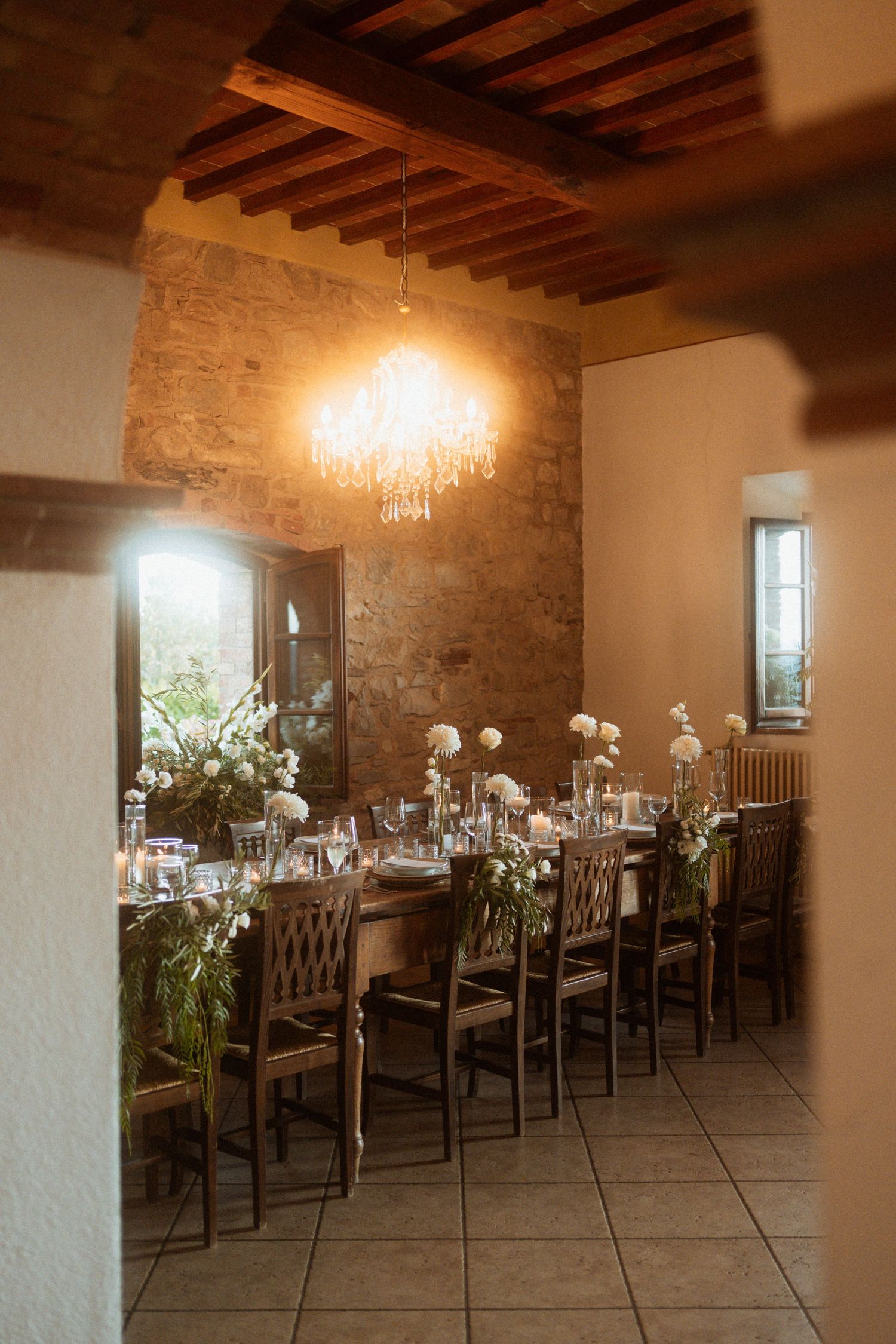 Elegant rustic dining room with long wooden table, floral centerpieces, crystal chandelier, and stone walls for an intimate event.