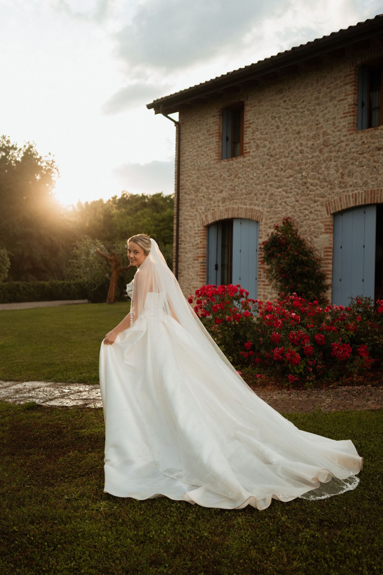 Bride in elegant white gown with long veil poses at golden hour outside rustic Italian villa with red roses.