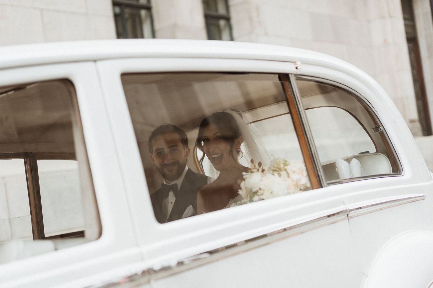 A bride and groom sitting together in a classic white vintage wedding car, smiling through the window.