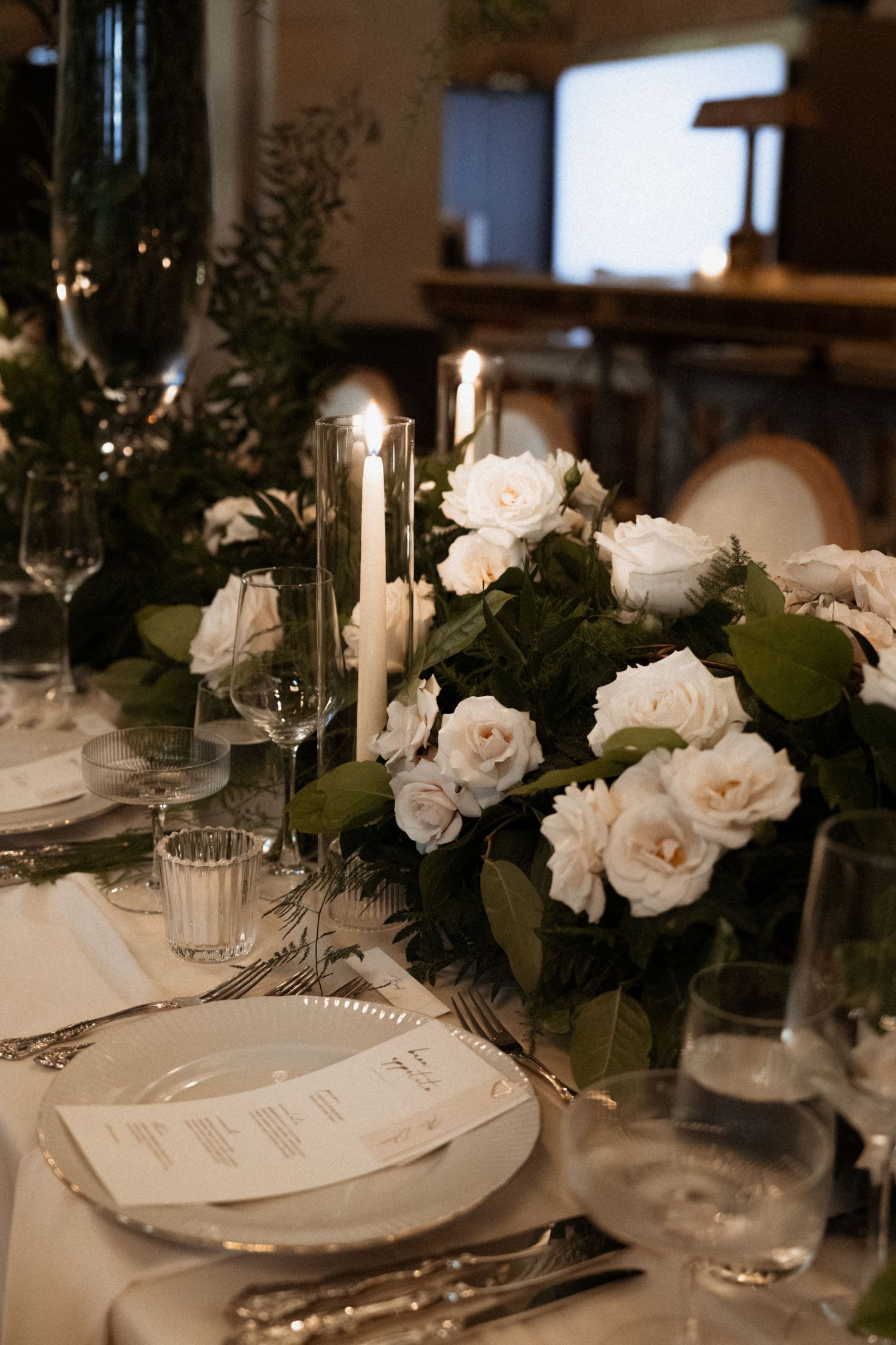Elegant wedding table setting with white roses, greenery centerpiece, candles, and silver charger plates.