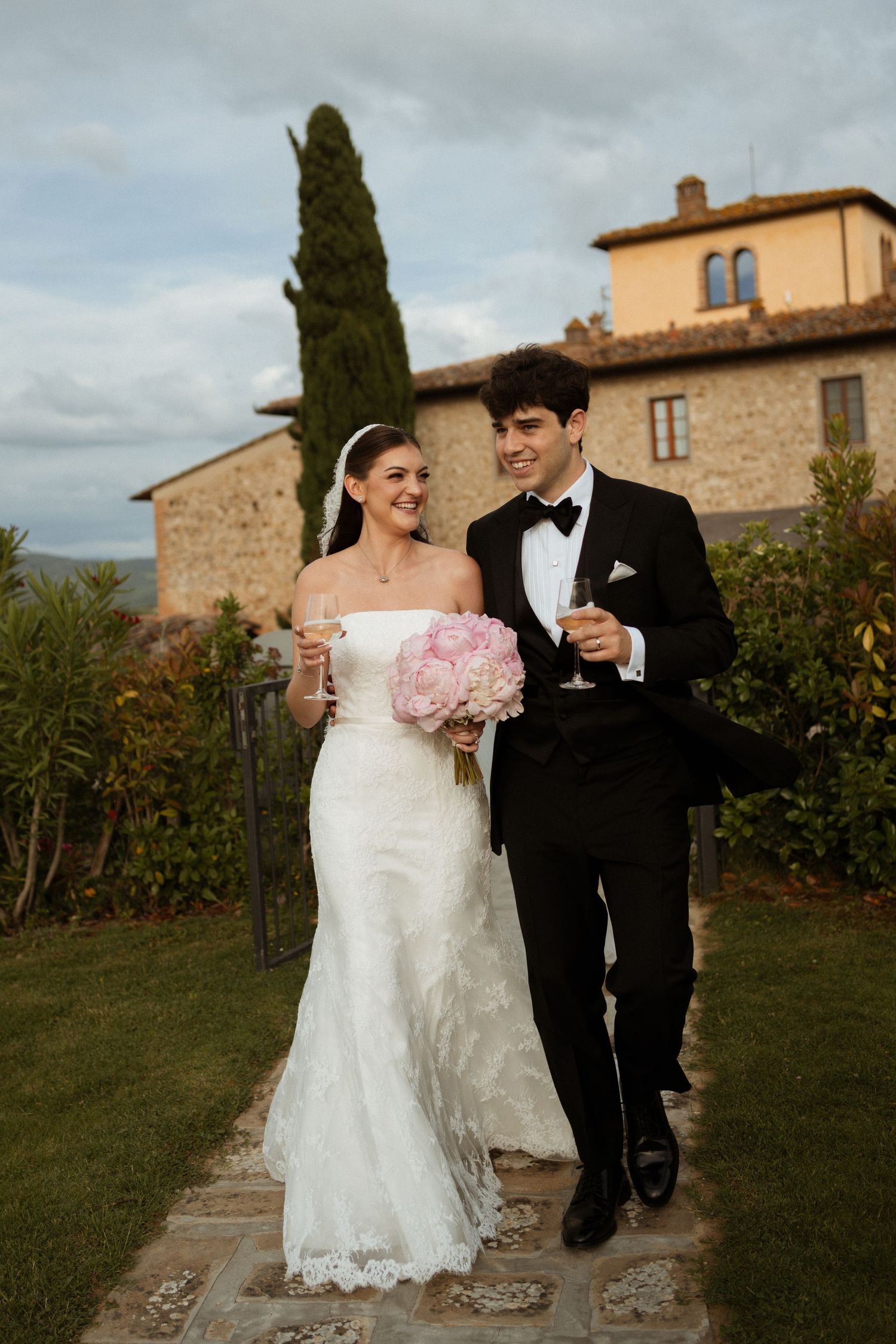 A bride and groom walk together outdoors at a Tuscan villa, holding champagne glasses and a pink peony bouquet.