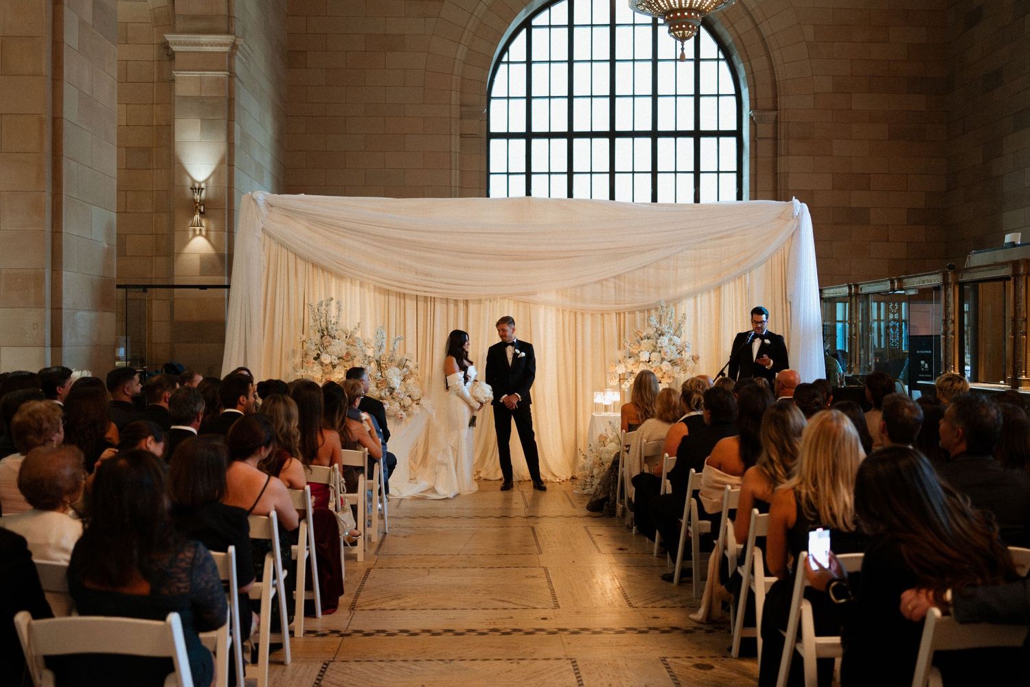 Wedding ceremony in an elegant historic venue with high windows and guests seated in rows for the service.