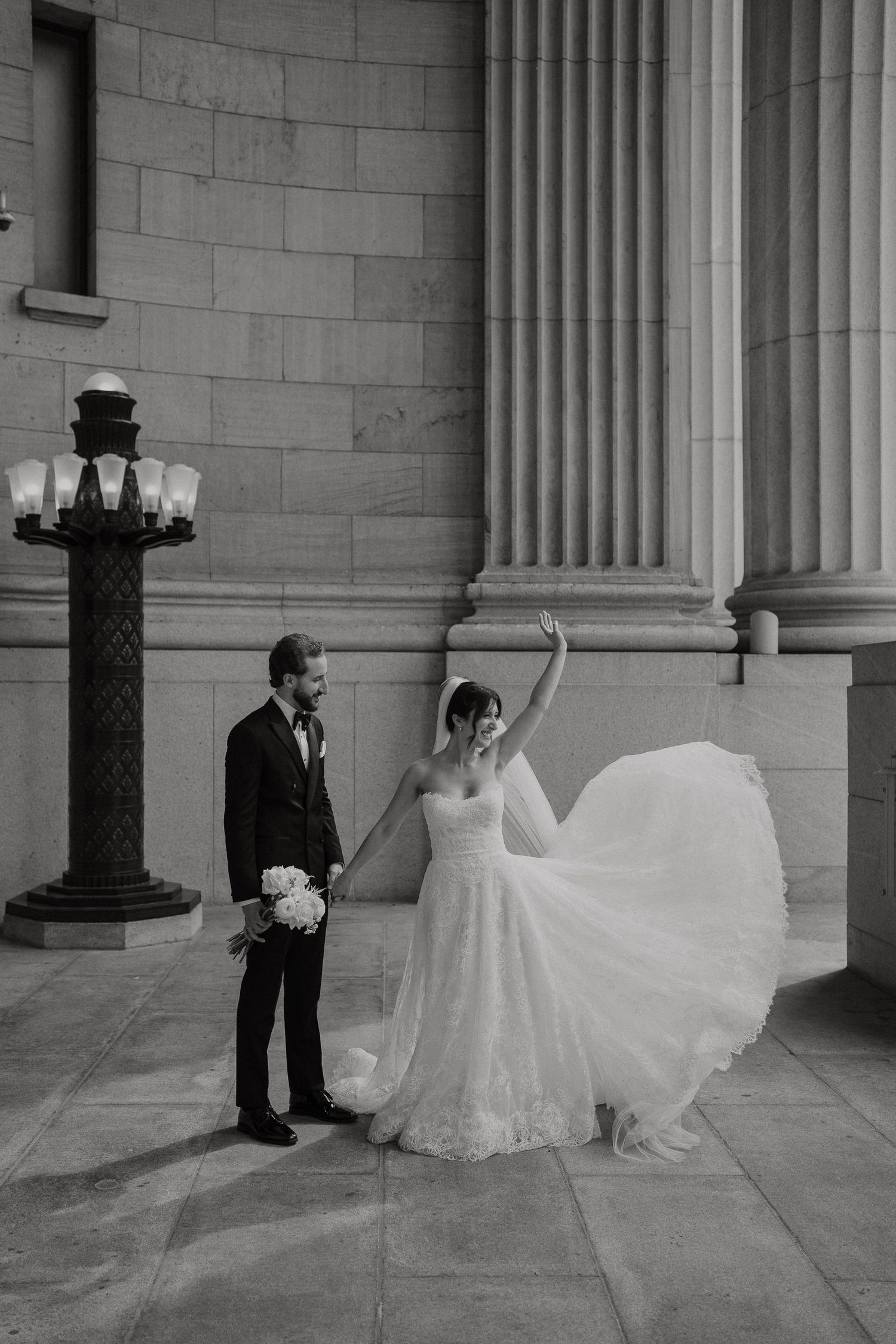 Black and white photo of bride twirling wedding dress beside groom holding bouquet near grand stone columns.