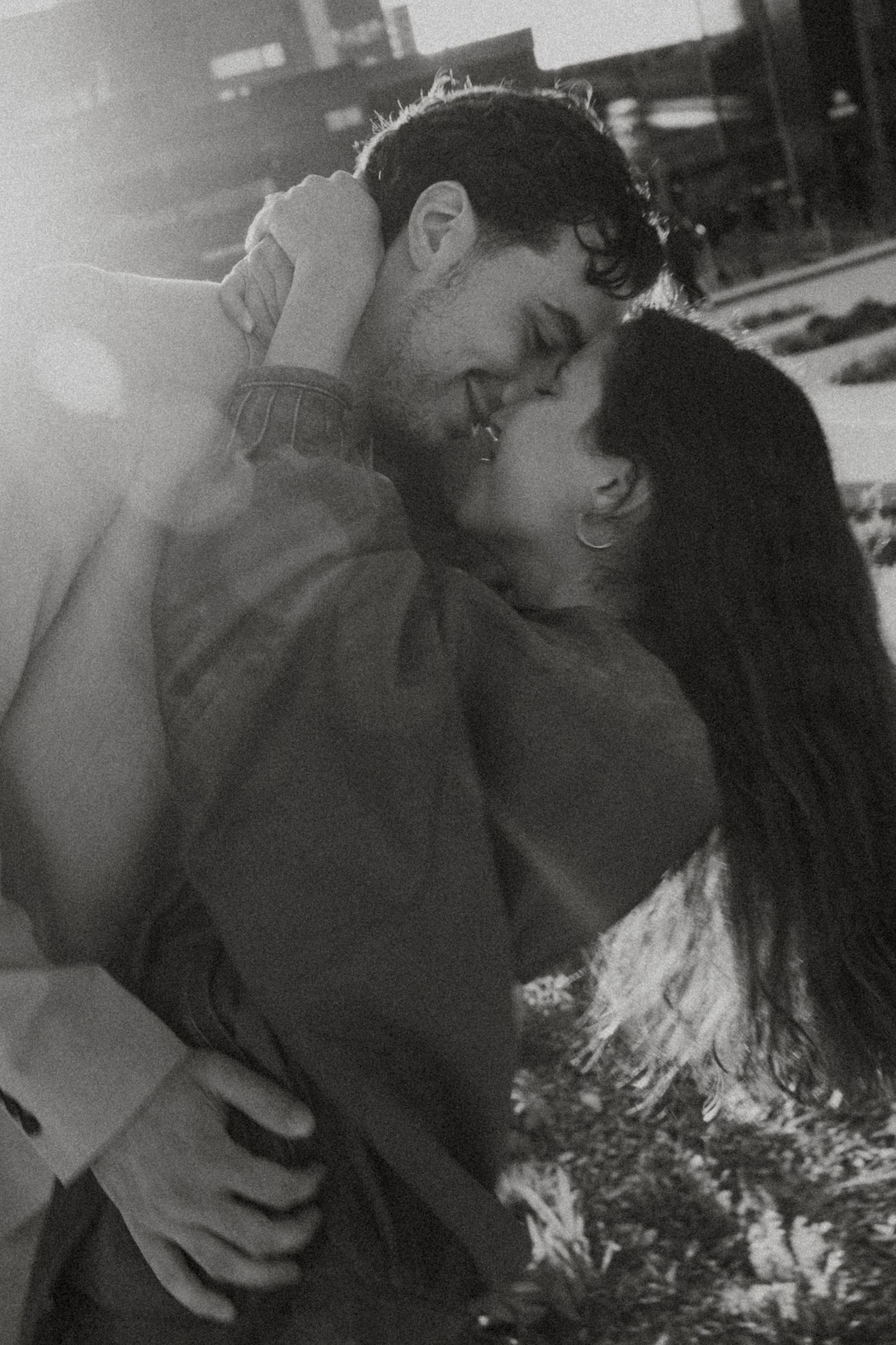 Black and white photo of a romantic couple sharing a tender kiss outdoors in warm backlit sunlight.