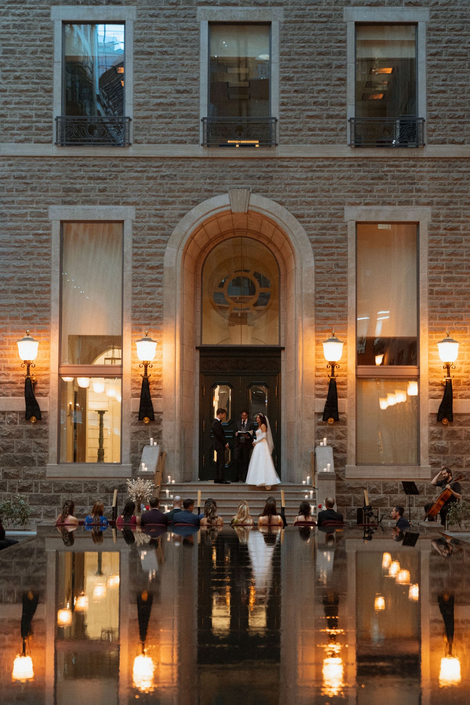 Illuminated lanterns reflect on a still pool outside a stone building with arched windows at dusk.