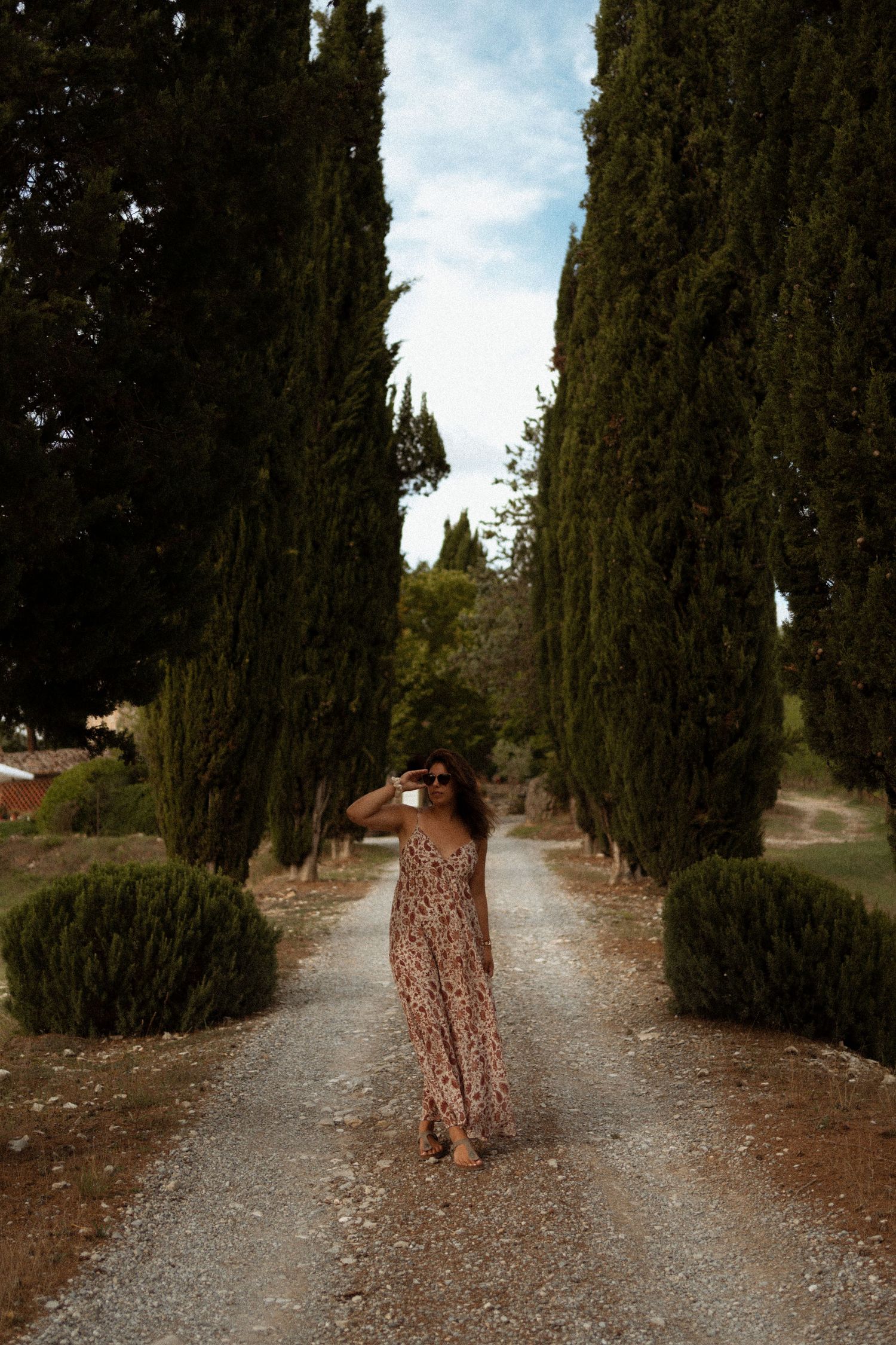 A long gravel path lined with tall cypress trees and manicured bushes leads through a scenic garden setting.