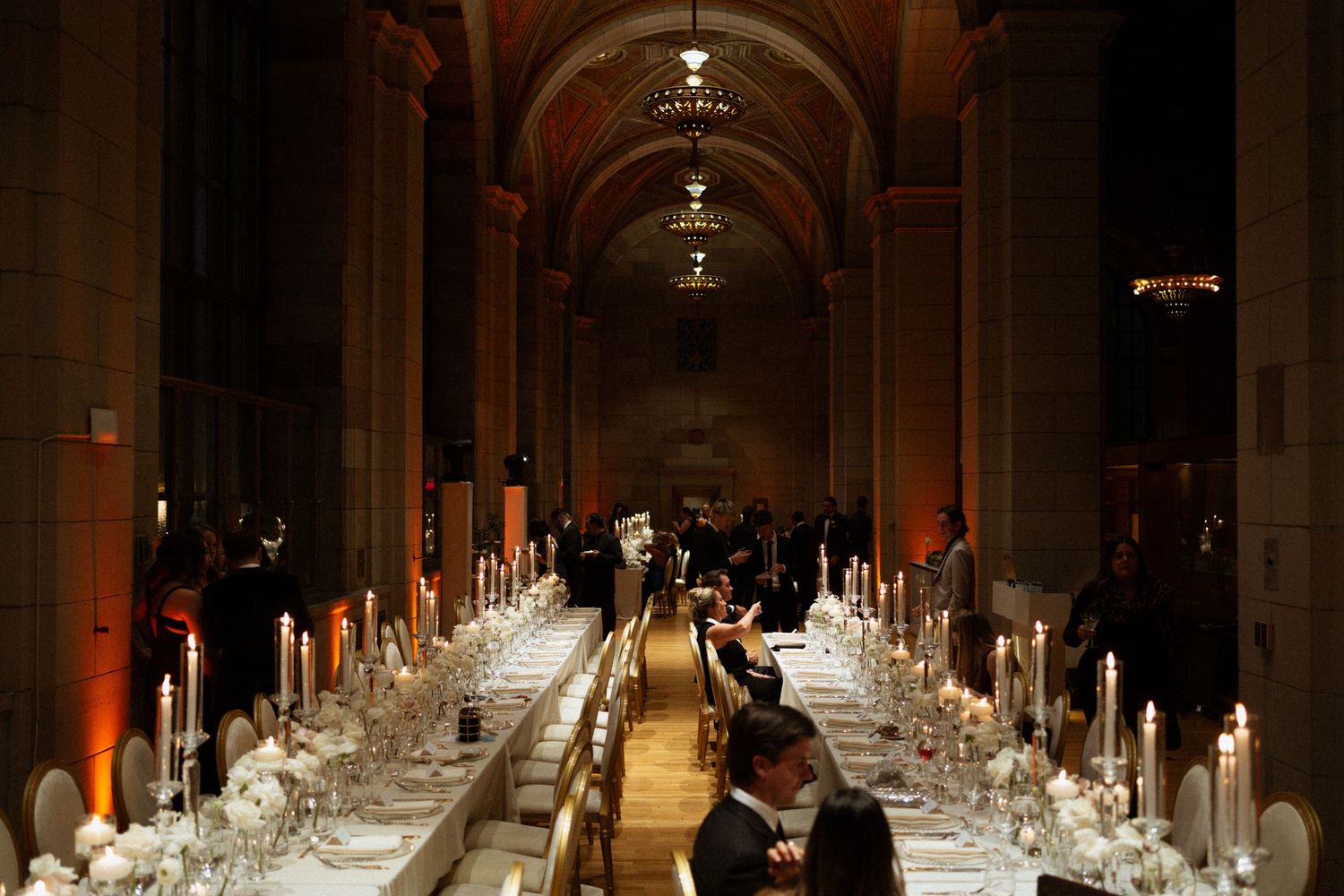 A long elegant banquet hall with multiple tables set for a formal dinner, illuminated by warm ambient lighting.